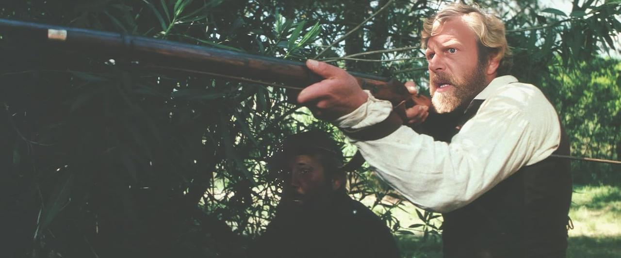 Ein Mann zielt mit einem Gewehr durch das Laub. Ein anderer Mann ist teilweise hinter ihm sichtbar. Grüne Bäume sind im Hintergrund.