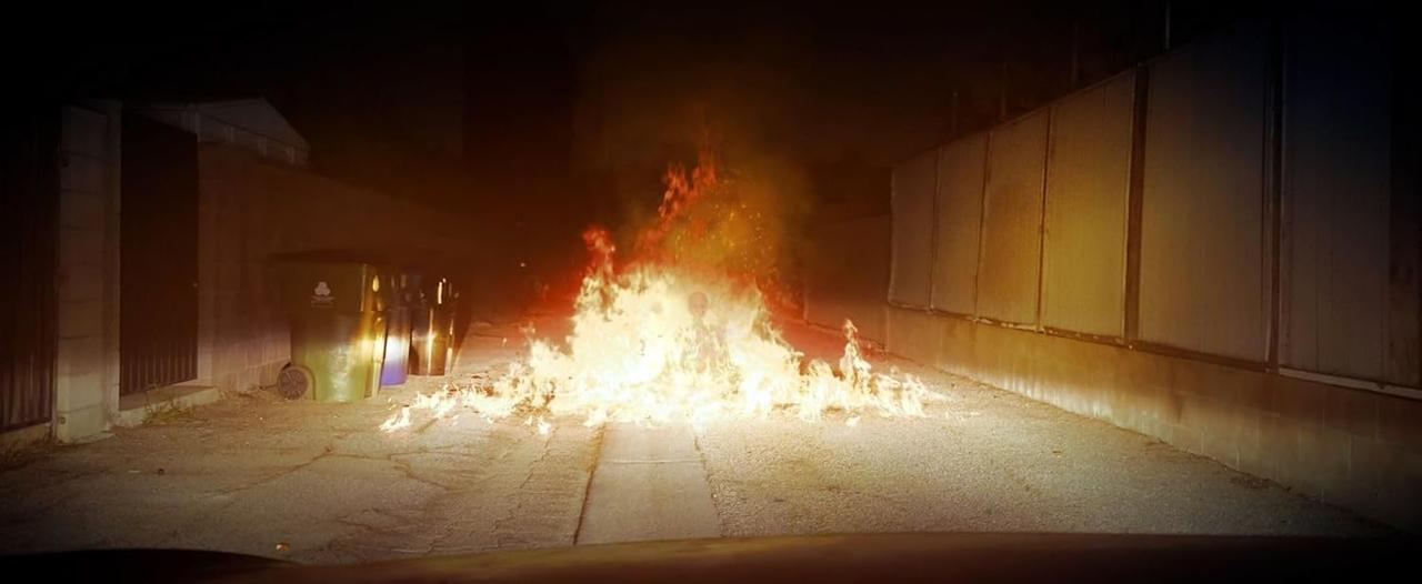 Ein Feuer brennt hell auf einer dunklen Straße und beleuchtet eine Mülltonne und eine Mauer. Flammen steigen hoch und werfen einen warmen Schein.