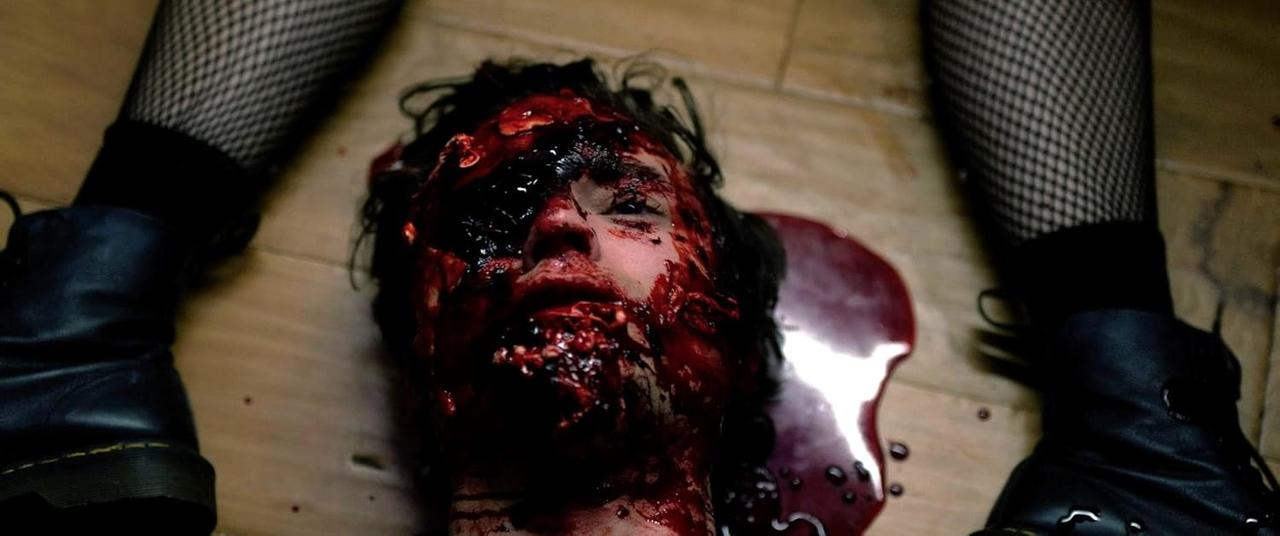 Ein blutiges Gesicht auf Holzboden, mit schwarzen Stiefeln und Netzstrümpfen. Blut um den Kopf.