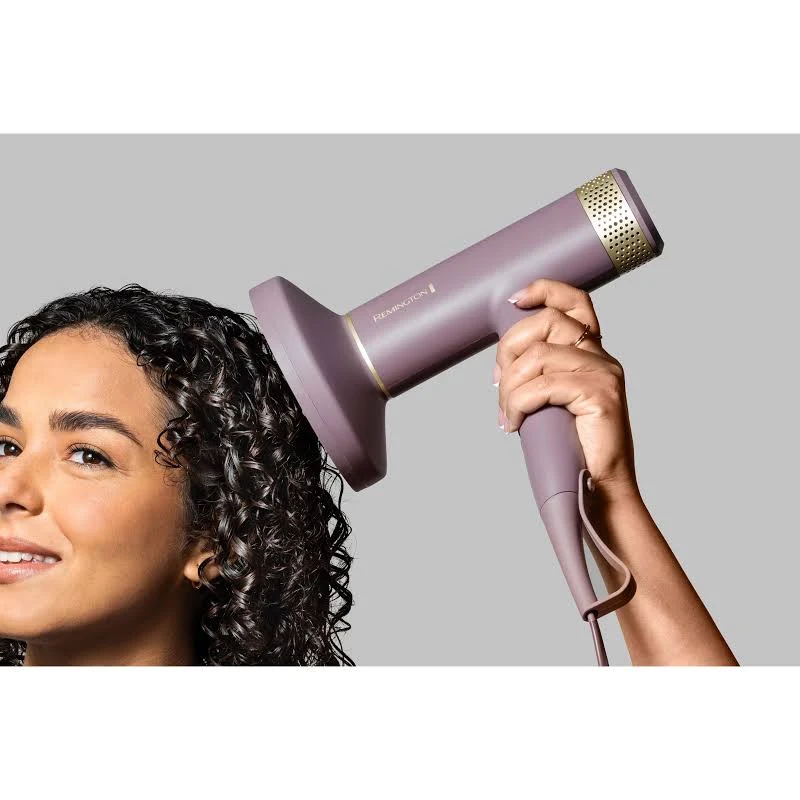 Una mujer usa un secador de pelo Remington morado en su cabello rizado. Sonríe, sosteniendo el secador con un acento dorado.