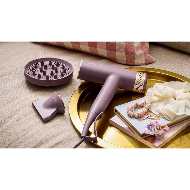 Un secador de pelo morado con accesorios, una bandeja dorada, coleteros y libros. Una almohada está al fondo.