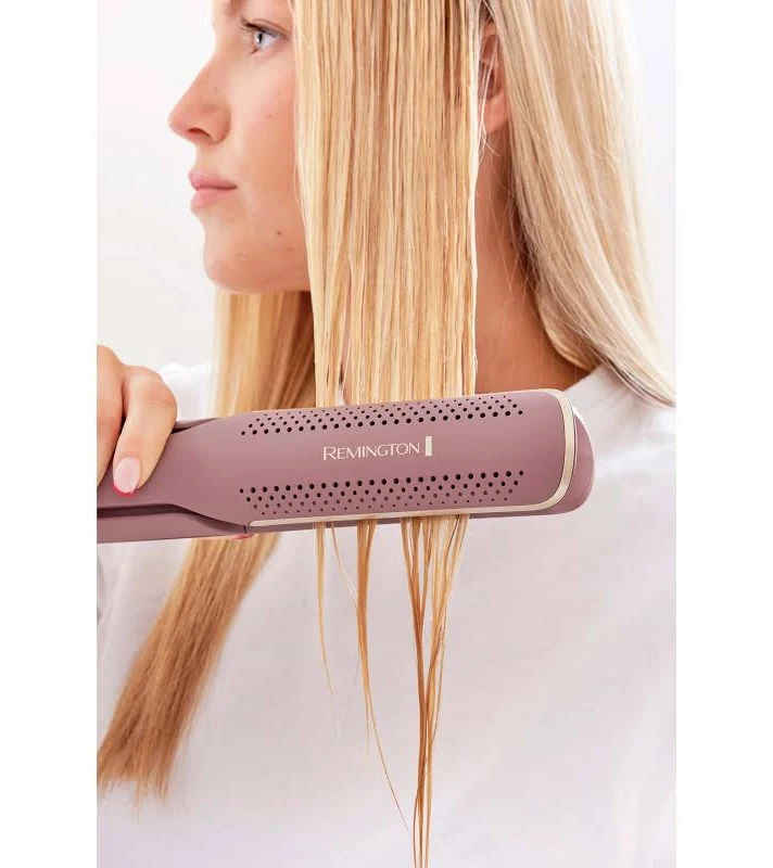 Una mujer se alisa su cabello rubio mojado con una plancha de pelo Remington. Viste una camisa blanca sobre un fondo blanco.