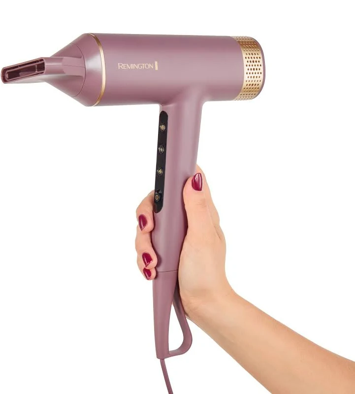 Una persona sostiene un secador de pelo Remington morado. El secador tiene detalles dorados. La persona tiene esmalte de uñas rojo.