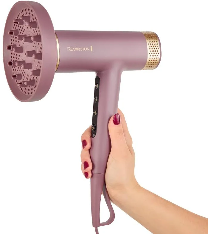 Una mano sostiene un secador de pelo Remington malva con difusor. El secador tiene detalles dorados y un panel de control negro.