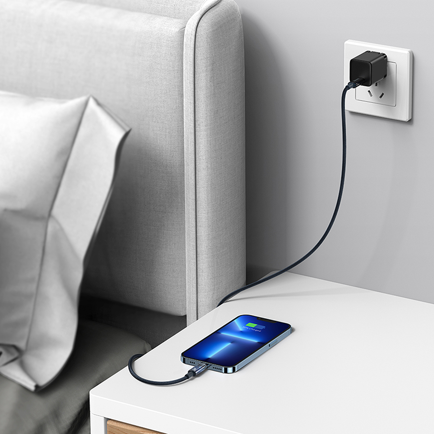 Un teléfono se está cargando en una mesita de noche blanca. Un cargador negro está enchufado a la pared y conectado al teléfono.