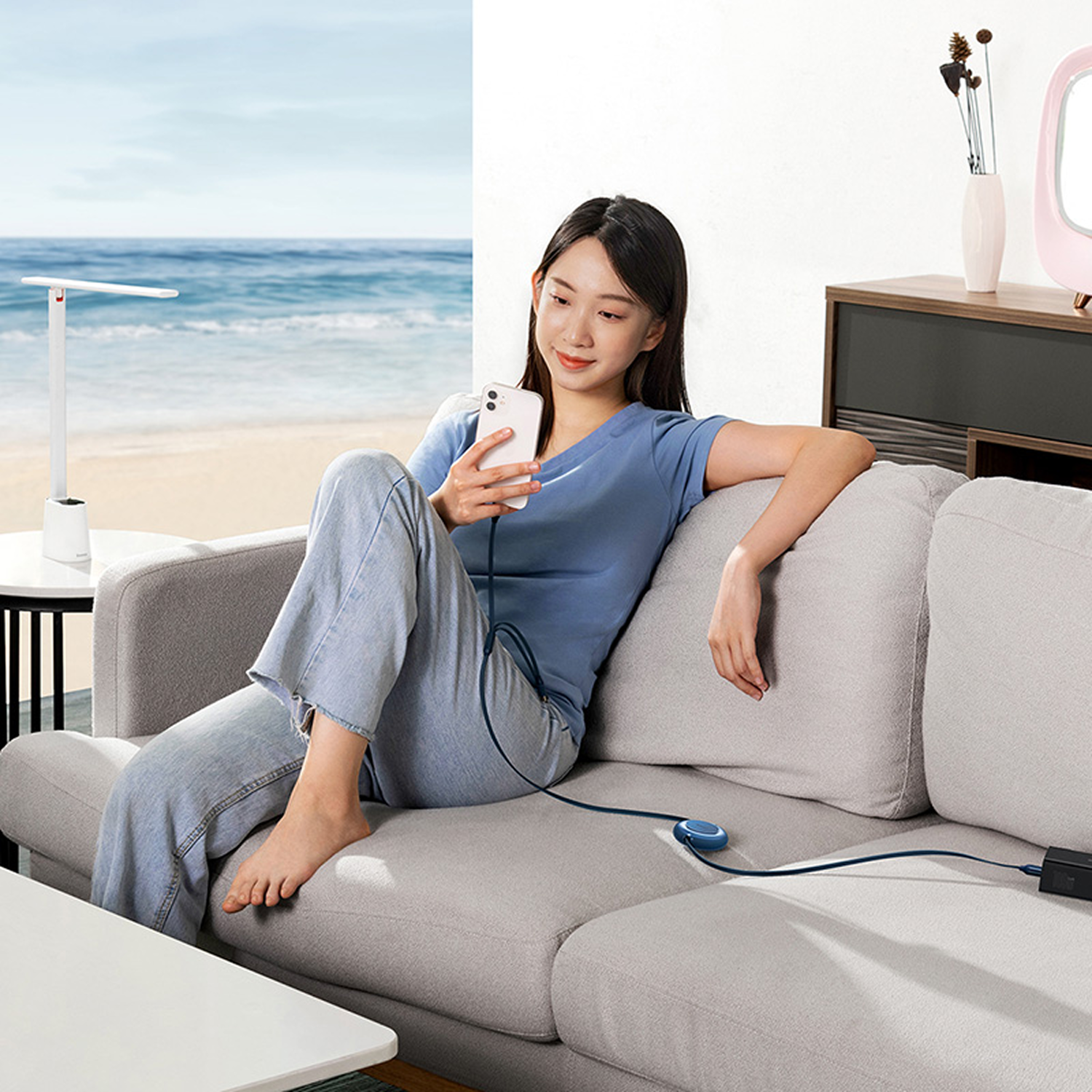 Mujer en sofá usando teléfono. Escena de playa visible. Lámpara y mesa auxiliar en el fondo.