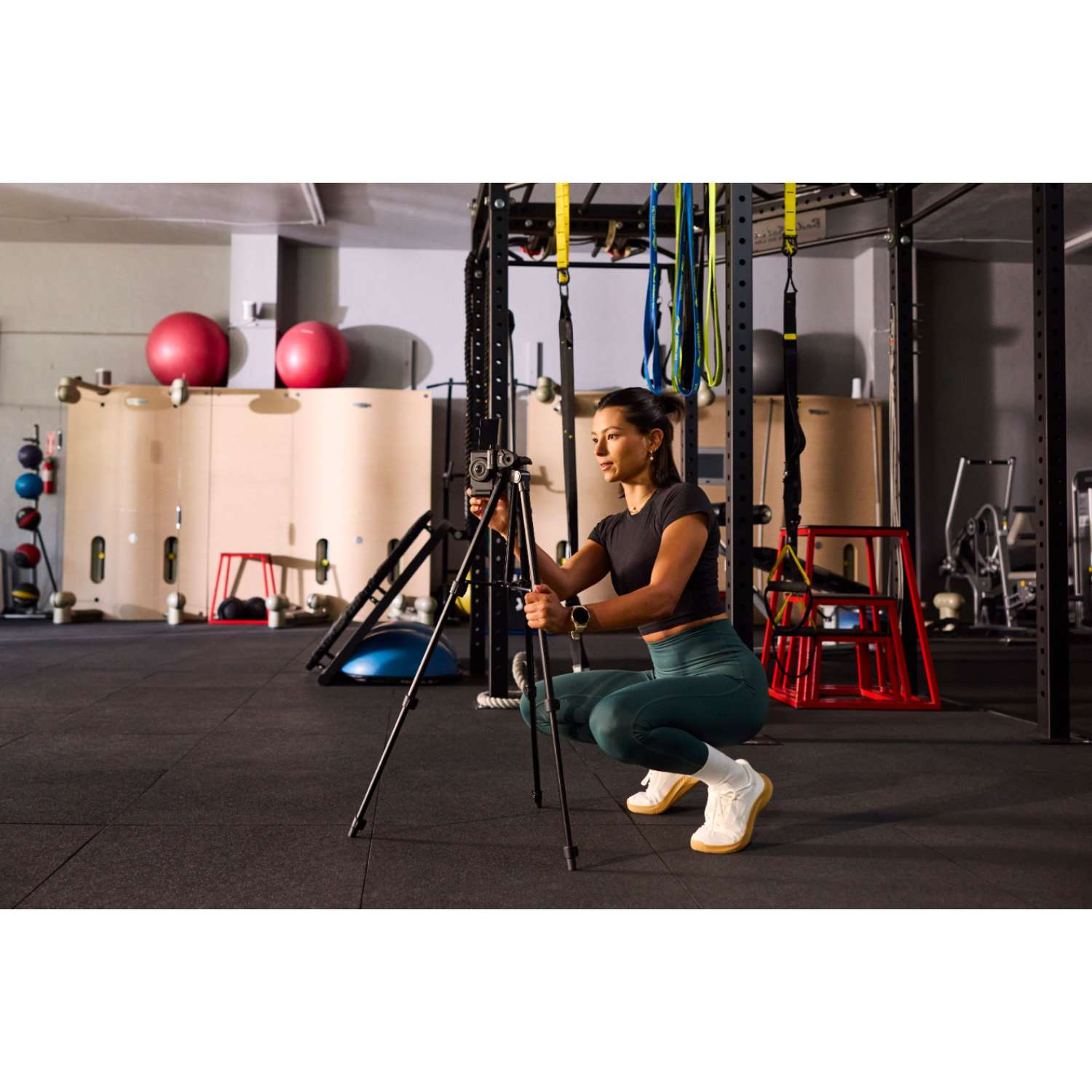 Mujer en el gimnasio montando una cámara en un trípode. Viste top negro, mallas verdes, zapatos blancos. Equipamiento de gimnasio.