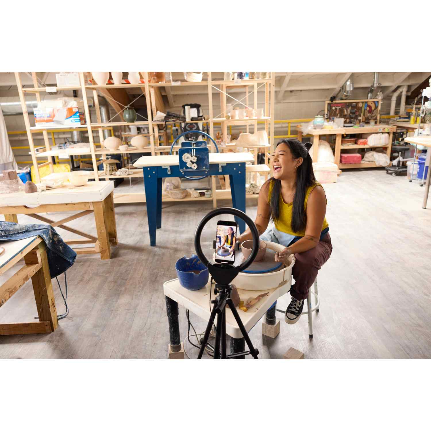 Mujer en taller ríe mientras hace cerámica. Teléfono en trípode la graba. Estanterías con cerámica y herramientas al fondo.