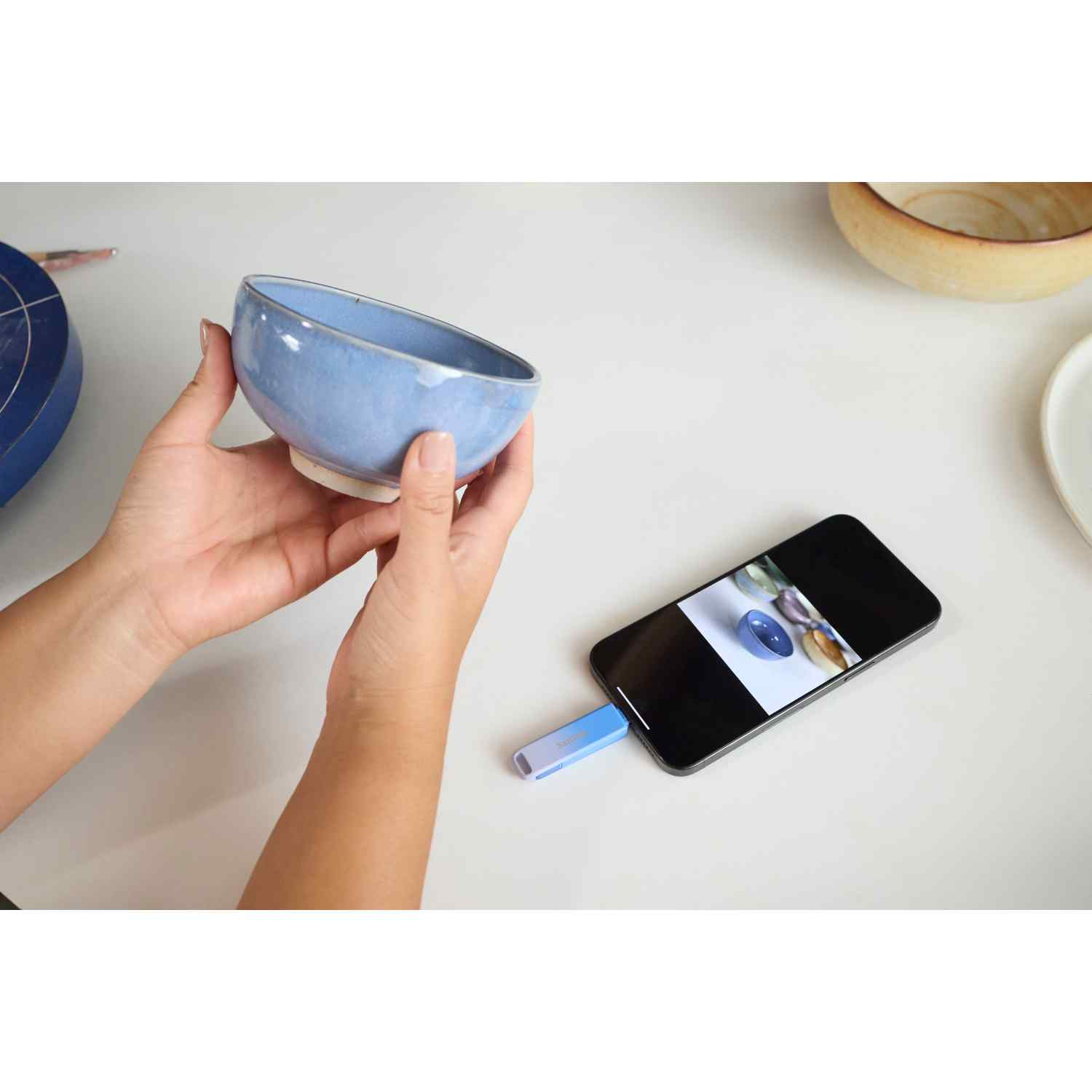 Manos sosteniendo un cuenco azul. Un teléfono muestra la imagen del cuenco. Otros cuencos y una unidad USB están en una mesa blanca.