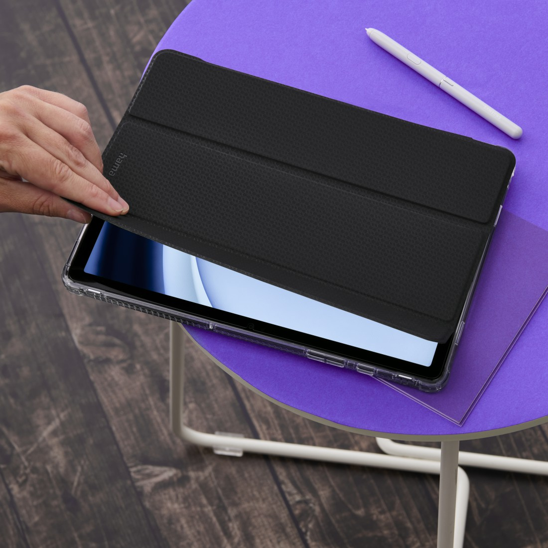 Eine Hand öffnet eine schwarze Tablethülle auf einem lila Tisch. Ein Stift und ein lila Blatt liegen ebenfalls auf dem Tisch. Holzboden im Hintergrund.