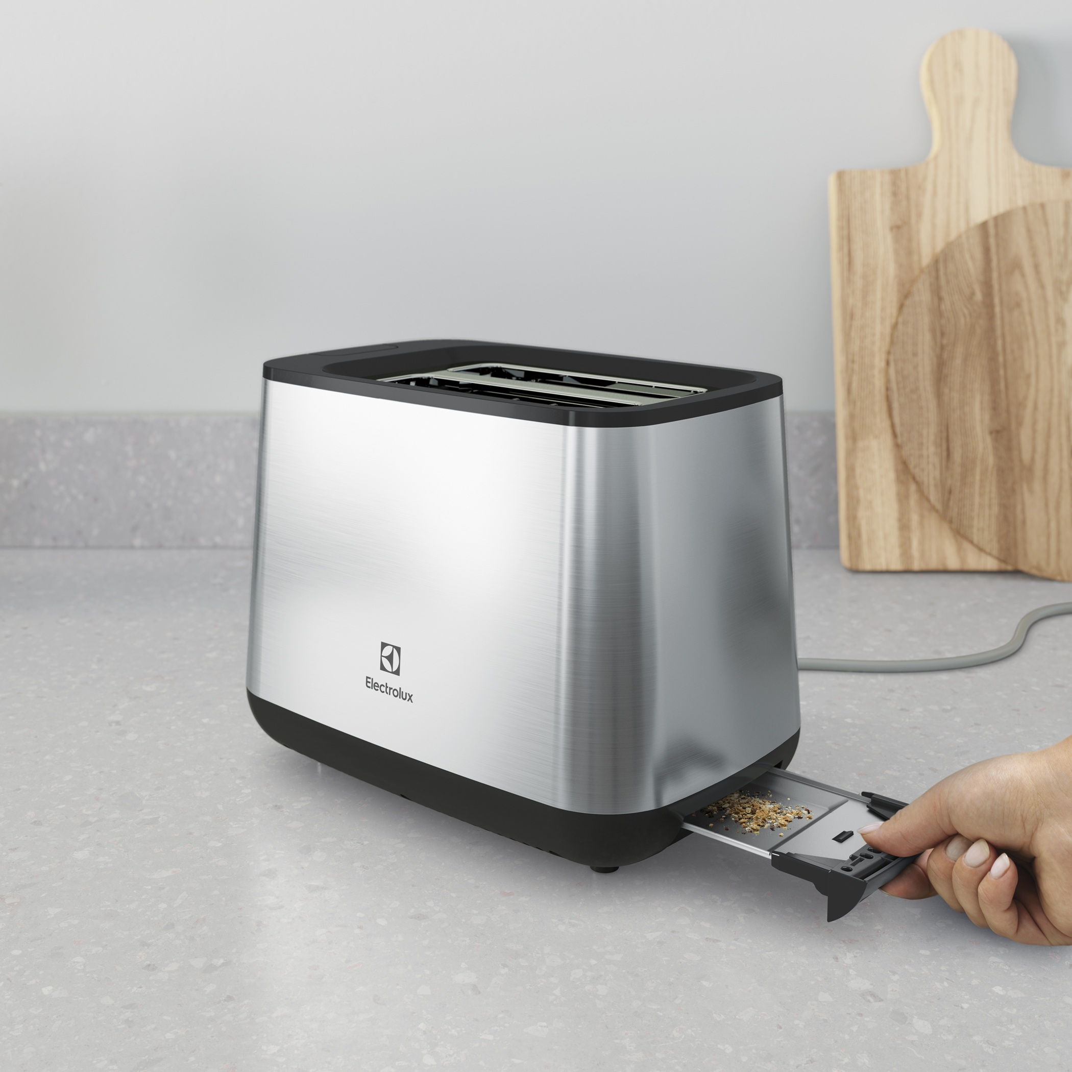 Gümüş renkli bir Electrolux tost makinesinde bir el kırıntı tepsisini çekiyor. Arka planda ahşap kesme tahtaları.