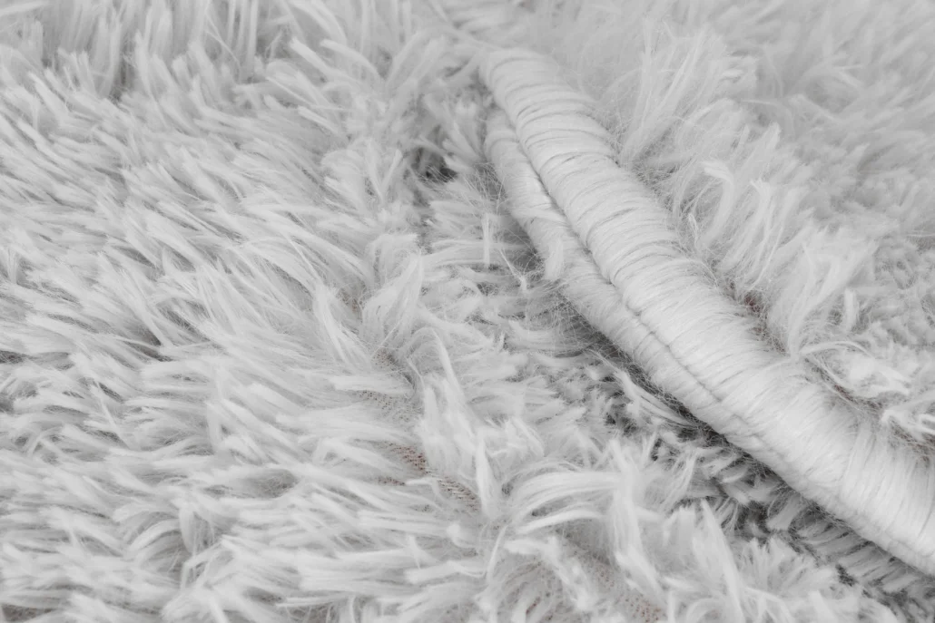 Zbliżenie białego, puszystego dywanu. Dywan ma długie, szorstkie włókna i zwiniętą krawędź.