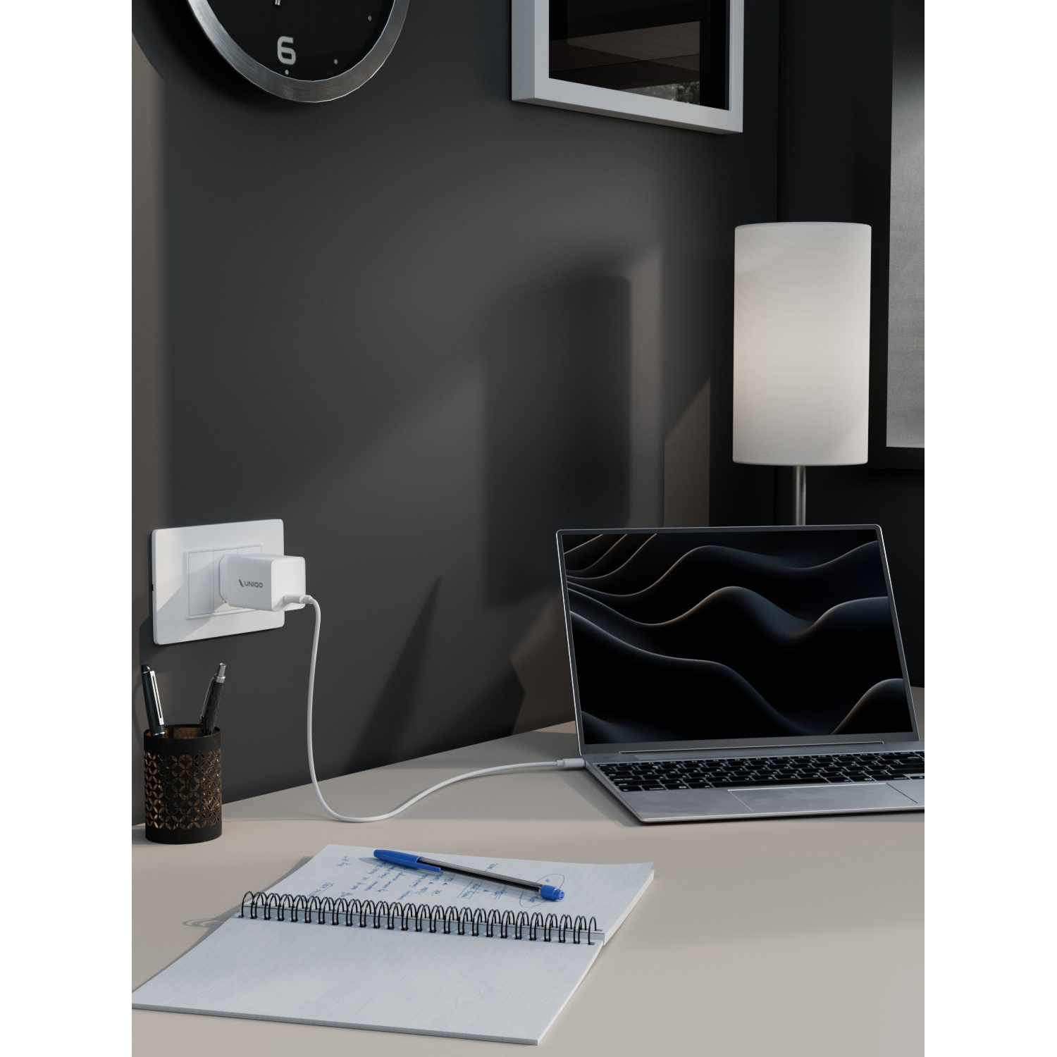 Laptop in carica sulla scrivania con taccuino, penna e lampada. Orologio da parete e presa con caricatore visibili. Muro grigio scuro.