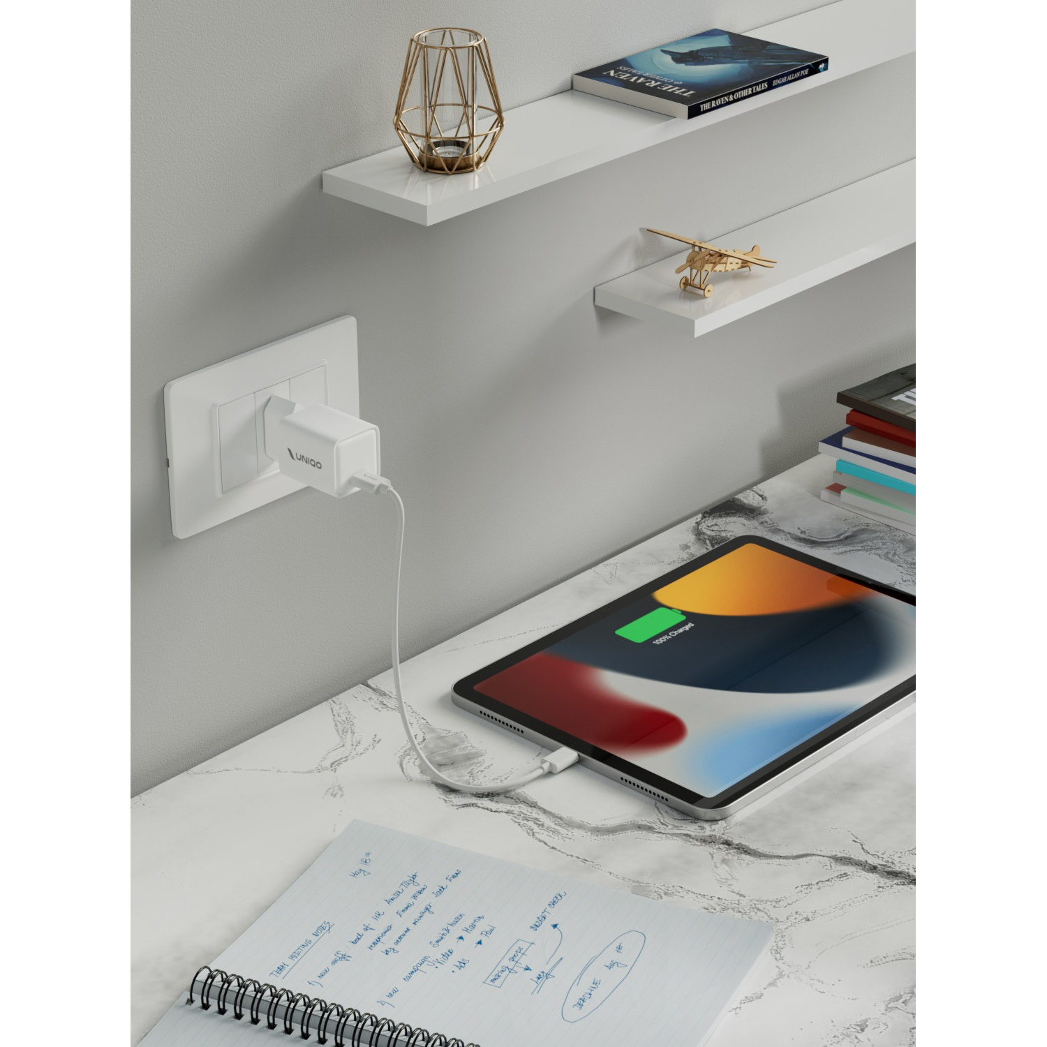 Tablet in carica su una scrivania in marmo. Taccuino con note, caricabatterie, mensole con decorazioni e un libro, e una presa sono visibili.