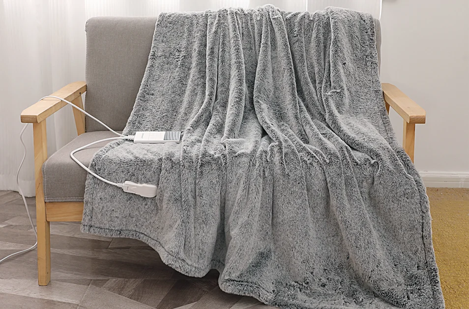 Szary koc grzewczy z imitacji futra jest narzucony na szary fotel. Koc ma panel sterowania i przewód zasilający.