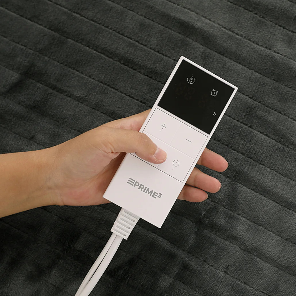 Ręka trzyma biały kontroler koca elektrycznego na tle ciemnoszarego, teksturowanego koca. Kontroler ma przyciski i wyświetlacz.