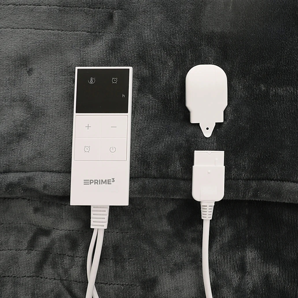 Biały kontroler i złącze koca elektrycznego na szarym kocu. Zawiera kontroler z przyciskami i wyświetlaczem oraz złącze.