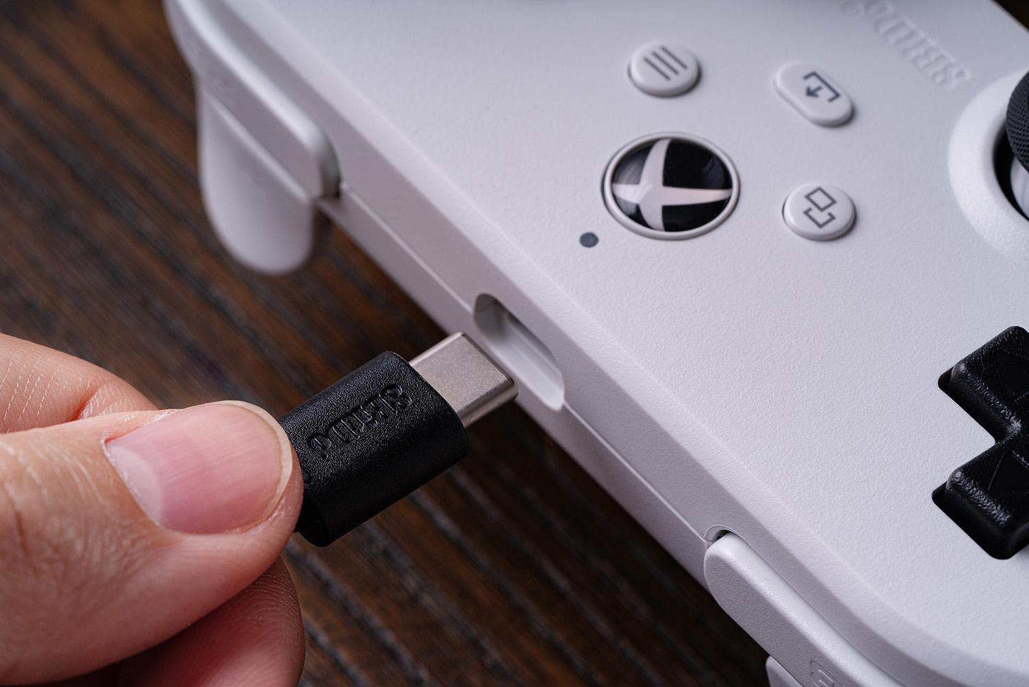 Eine Hand steckt ein schwarzes USB-C-Kabel in einen weißen Gamecontroller. Der Controller liegt auf einer Holzoberfläche.