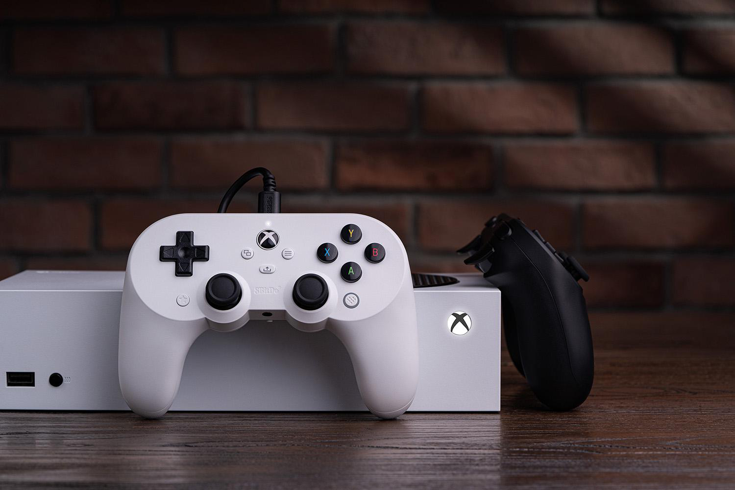 Xbox Series S Konsole mit weißen und schwarzen Controllern auf Holz vor einer Backsteinmauer.