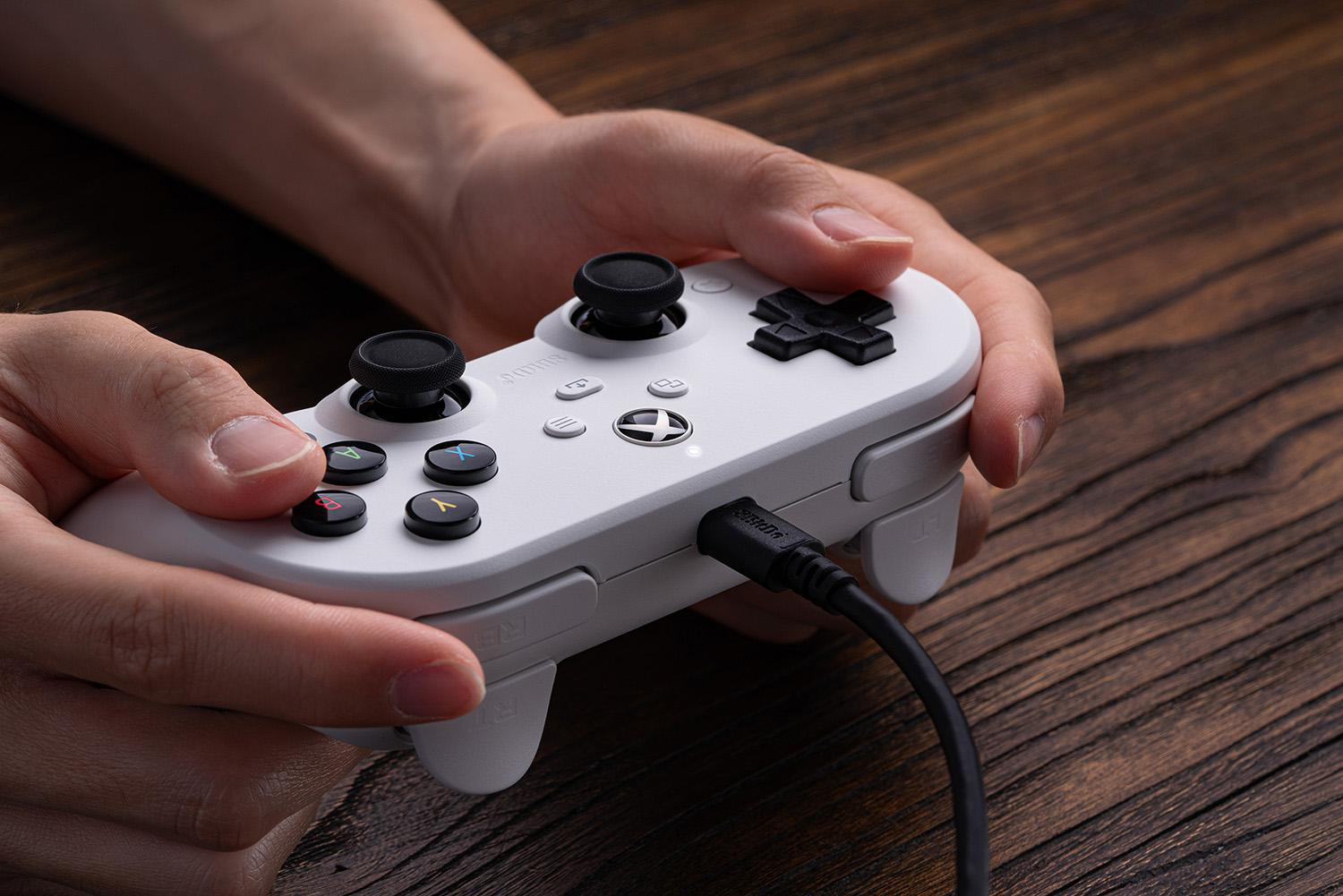 Hände halten einen weißen Gamecontroller mit schwarzen Knöpfen. Ein schwarzes Kabel ist eingesteckt. Holz Hintergrund.