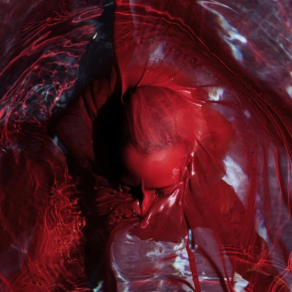 Ein Kopf, der in roter Flüssigkeit, möglicherweise Farbe, untergetaucht ist. Die Flüssigkeit hat Kräuselungen und Falten.