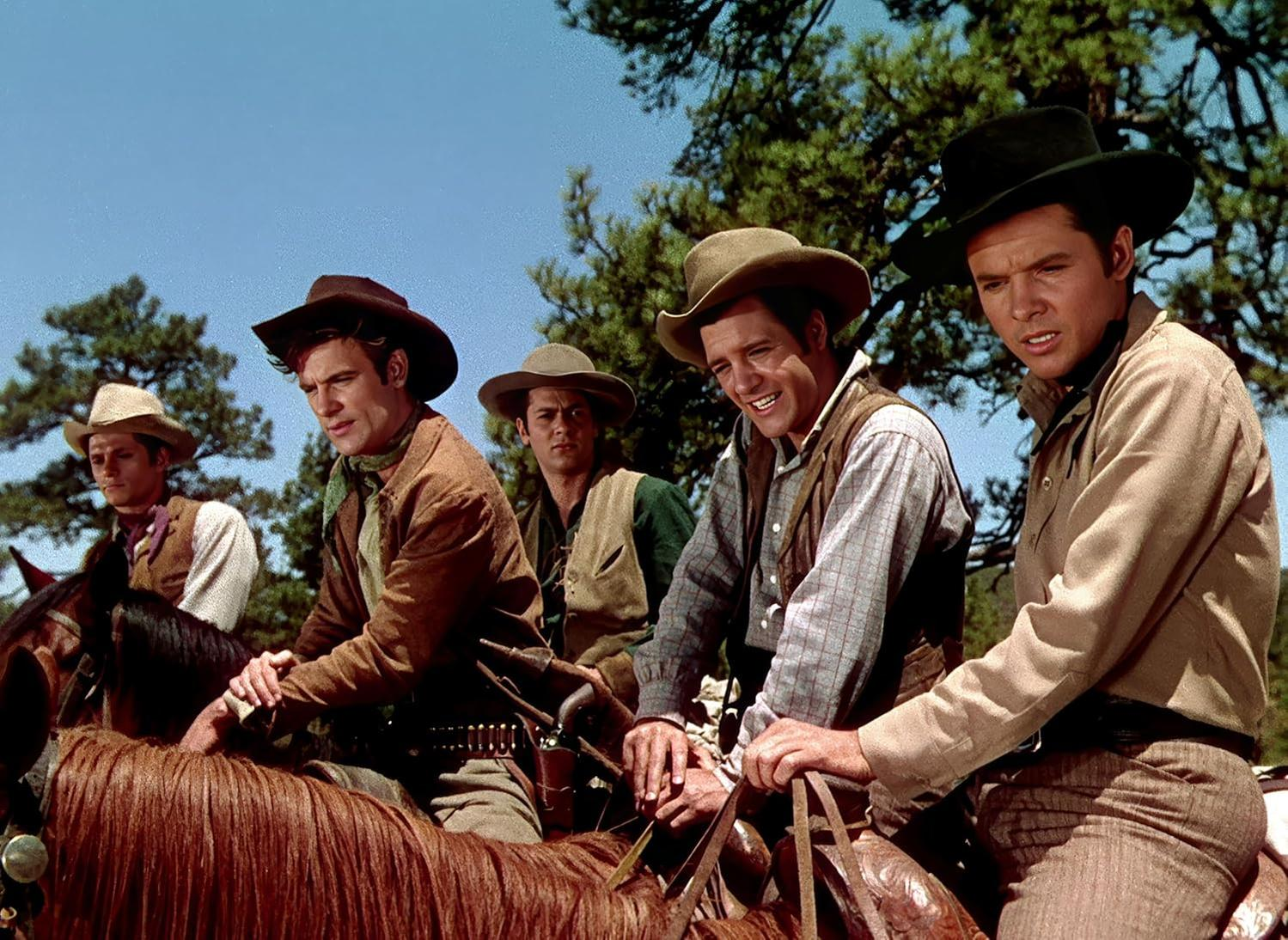 Sechs Cowboys auf Pferden, die Hüte und Westen tragen, vor blauem Himmel. Bäume sind im Hintergrund. Sie lächeln.