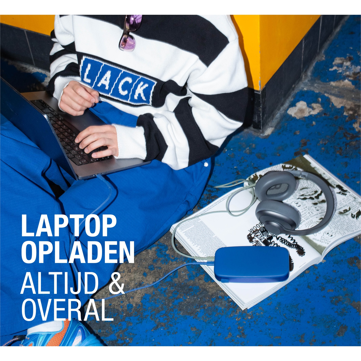 Persoon met laptop, verbonden met blauwe powerbank. Hoofdtelefoon en tijdschrift zijn zichtbaar. De persoon draagt ​​een blauwe overall en een gestreepte trui.