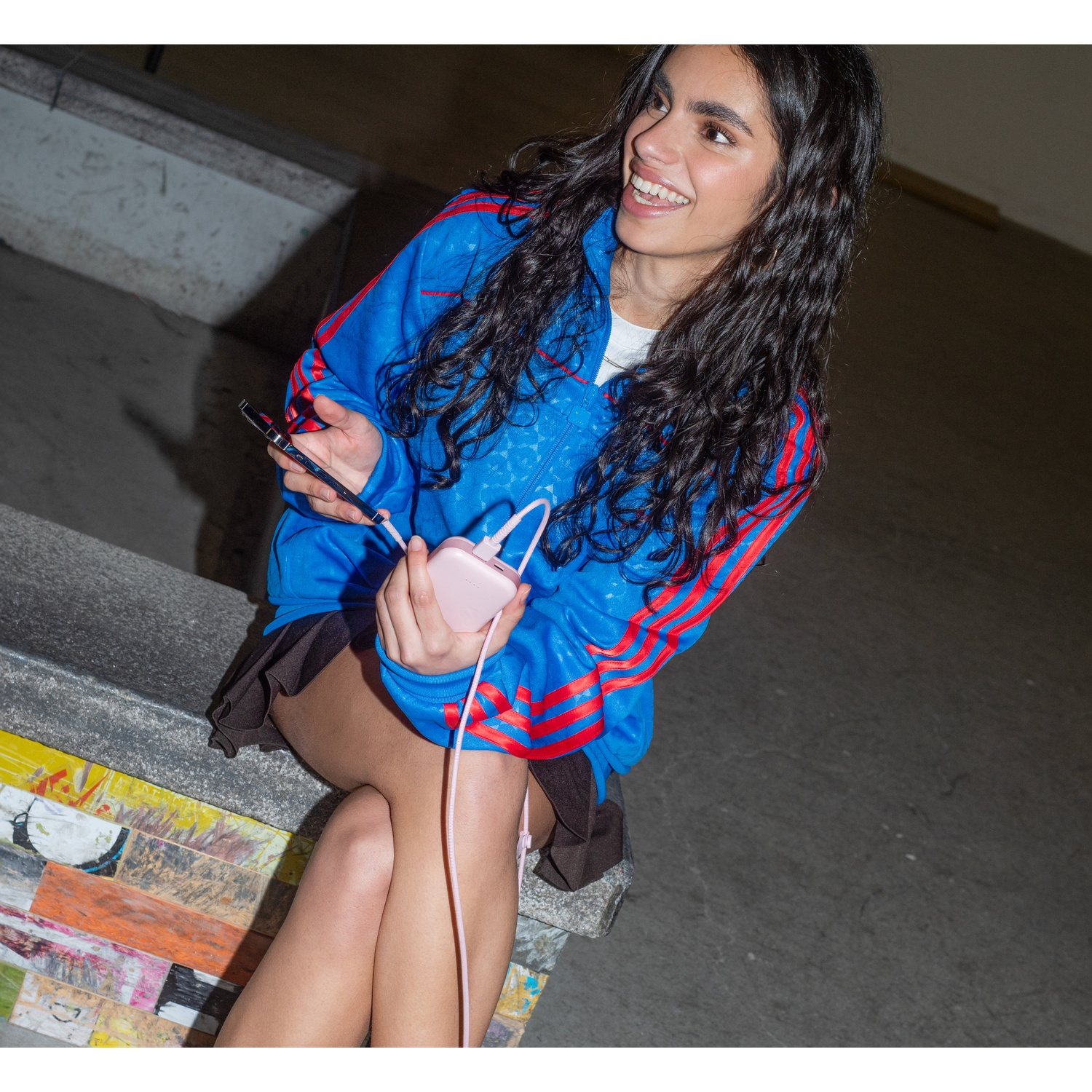 Une femme souriante en veste bleue tient un téléphone et une batterie externe rose. Elle est assise sur une structure en béton peinte de couleurs vives.