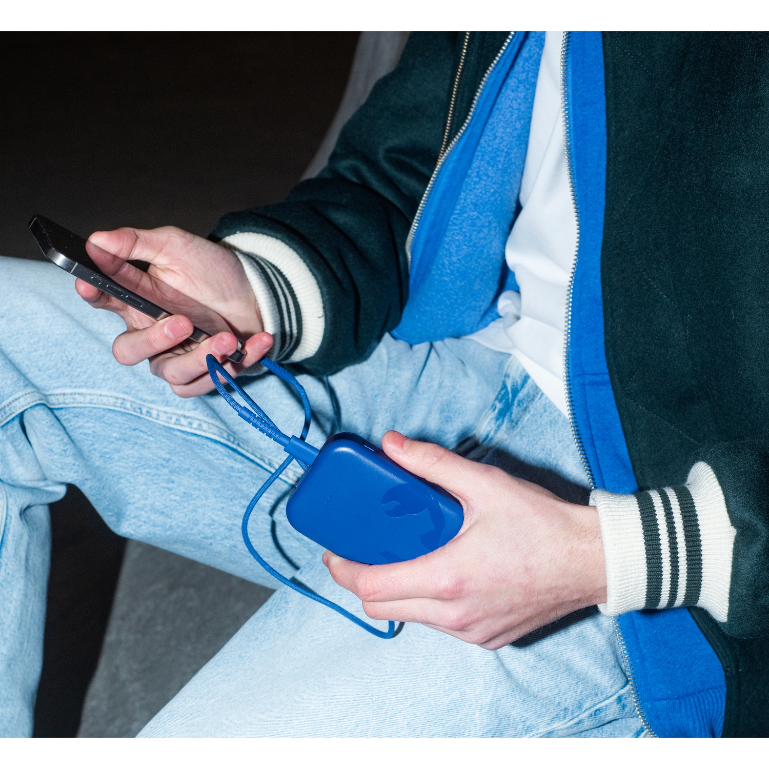 Une personne tient un téléphone et une batterie externe bleue. Elle porte une veste verte, une chemise bleue et un jean. Un câble bleu les relie.