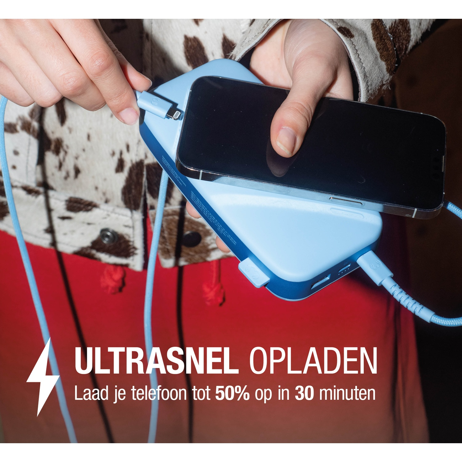 A person charging a black phone on a blue power bank. The person is wearing a cow-print jacket and red shirt.