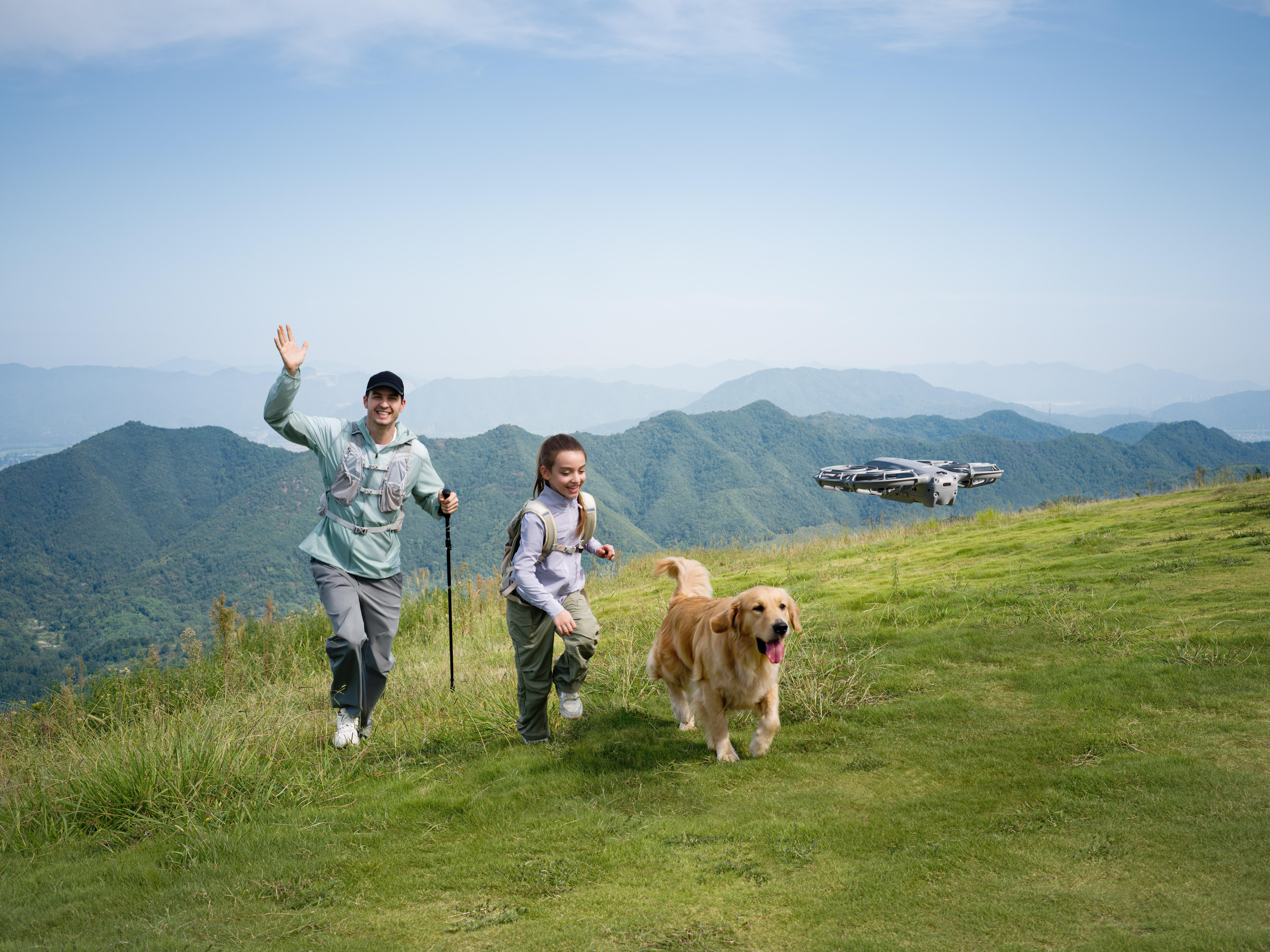 Ein Mann, ein Mädchen und ein Hund rennen auf einem grünen Hügel. Eine Drohne fliegt im Hintergrund. Berge und blauer Himmel.