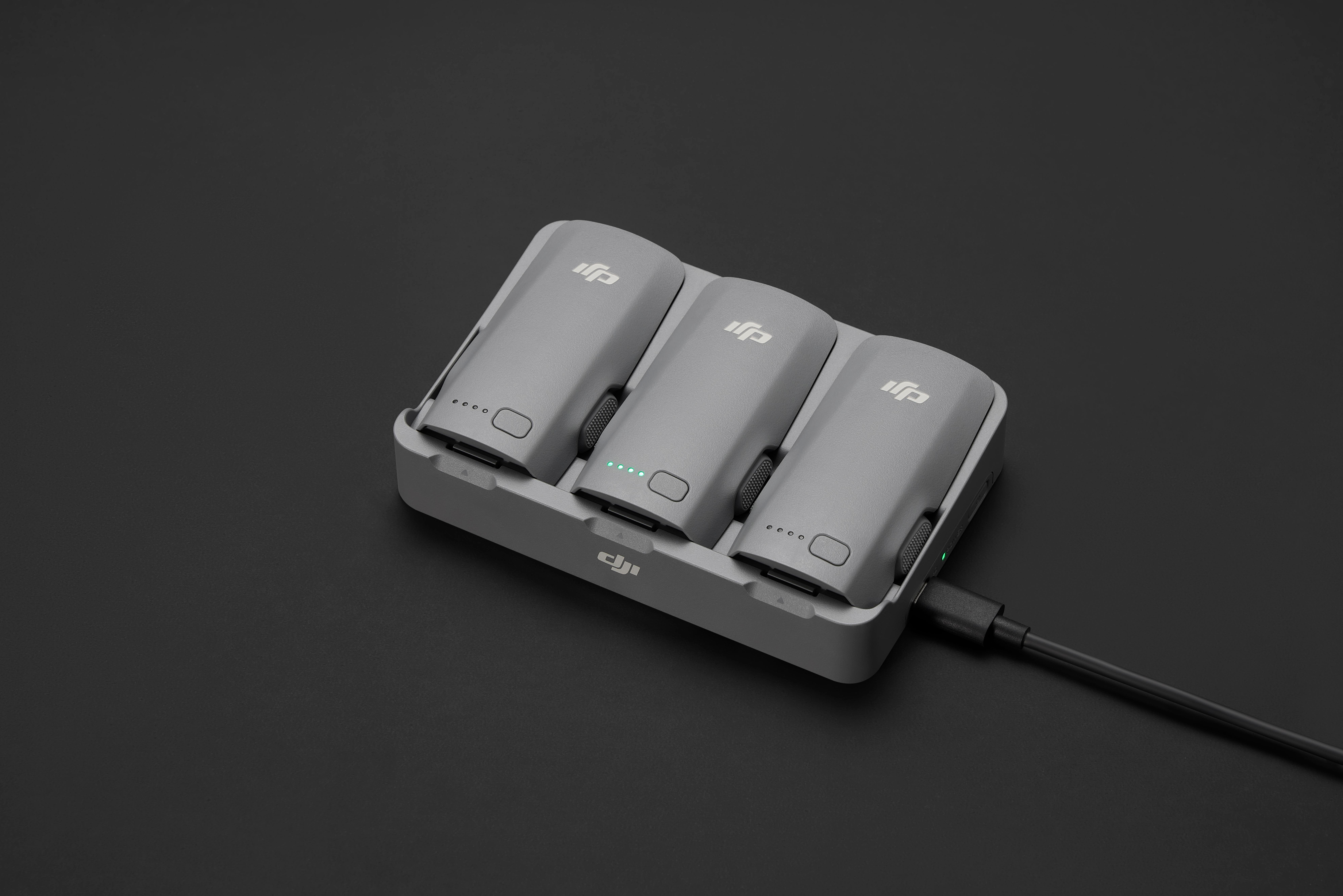Drei graue Drohnenbatterien in einer Ladestation auf schwarzer Oberfläche. Eine Batterie zeigt Ladelichter.