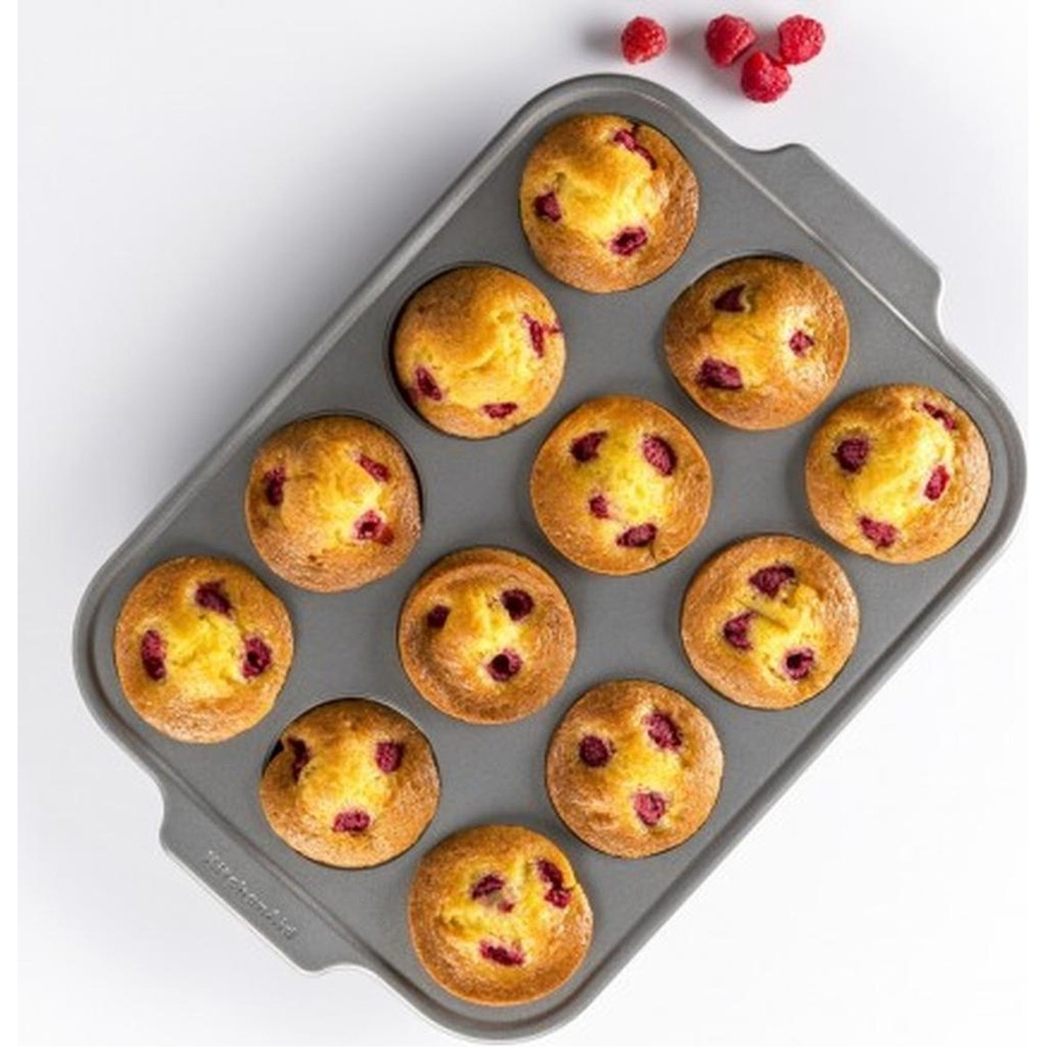 Een grijze muffinvorm bevat 12 muffins met frambozen. Drie frambozen liggen op het witte oppervlak.