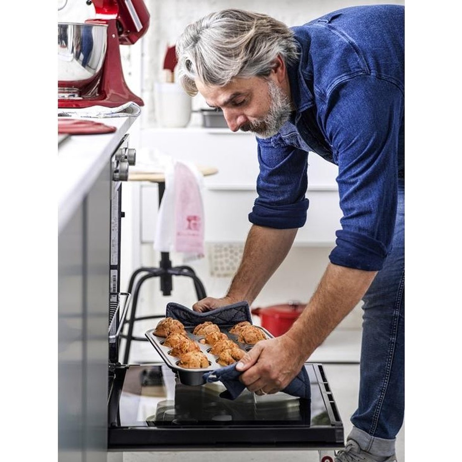 Een man in een blauw overhemd en spijkerbroek haalt een bakplaat met muffins uit de oven. Keukenapparatuur is zichtbaar op de achtergrond.