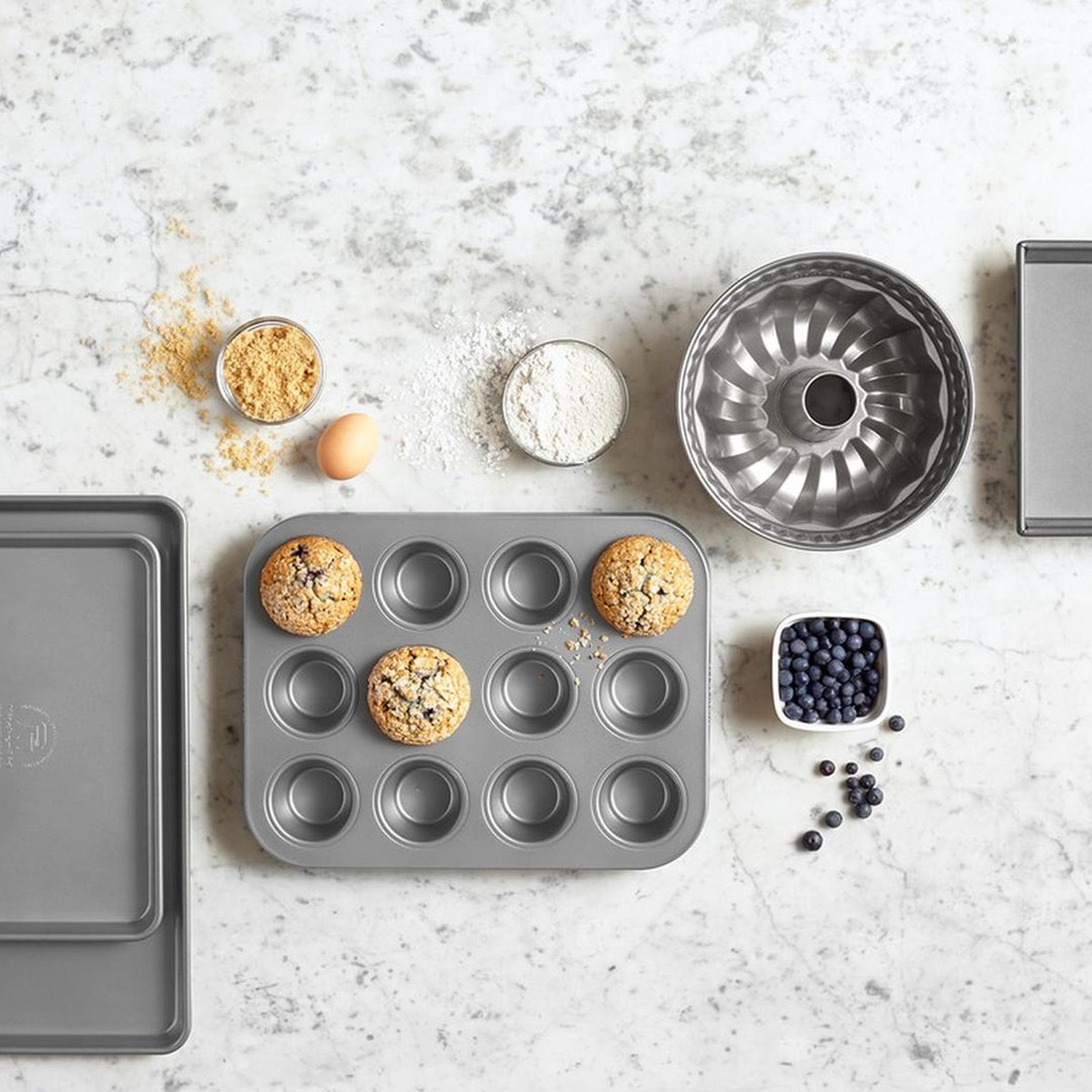 Bovenaanzicht van bakbenodigdheden: muffinvorm met muffins, kommen met bloem, bruine suiker en bosbessen, tulbandvorm, ei.