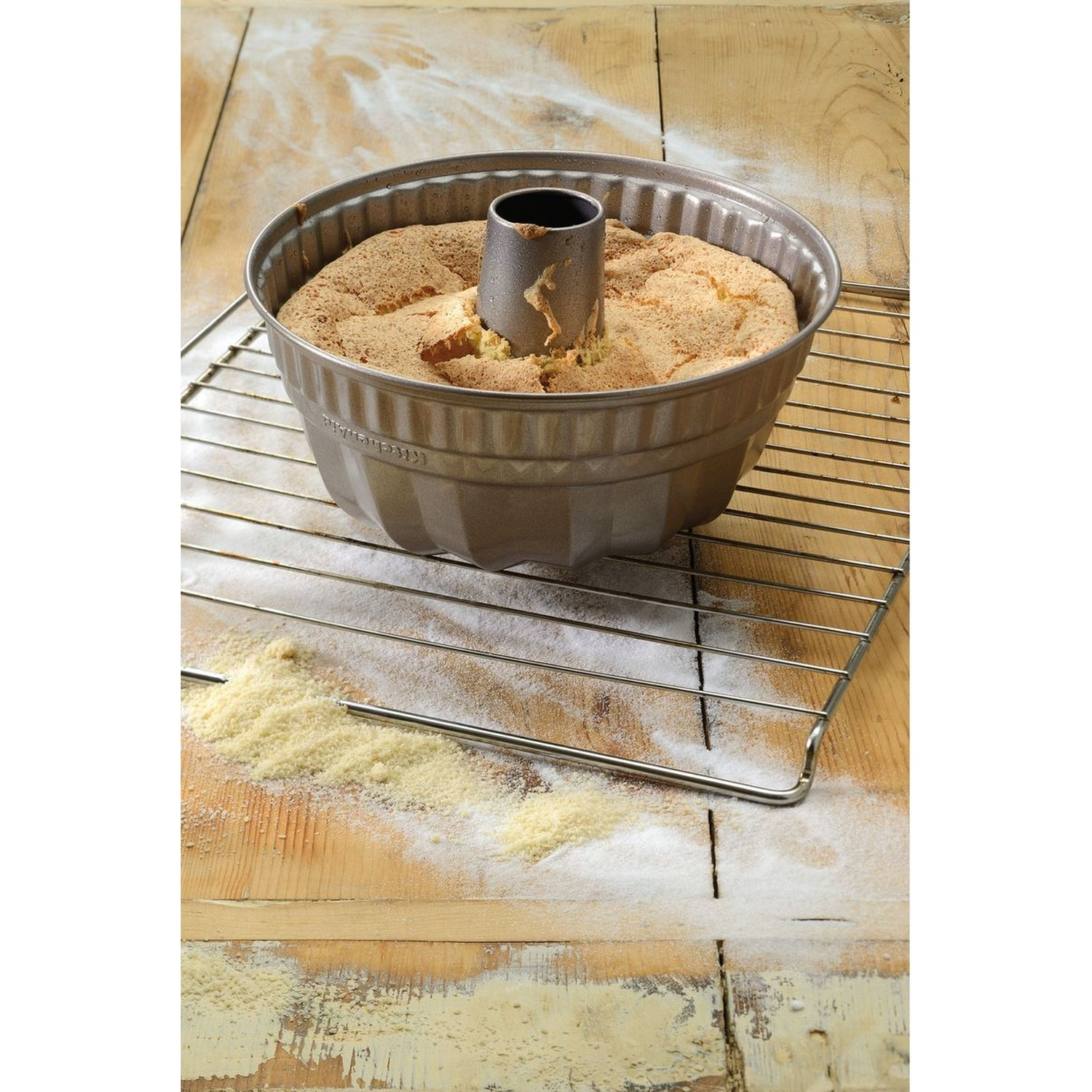 Un gâteau dans un moule à manqué est posé sur une grille sur une surface en bois saupoudrée de farine. Le gâteau est doré.