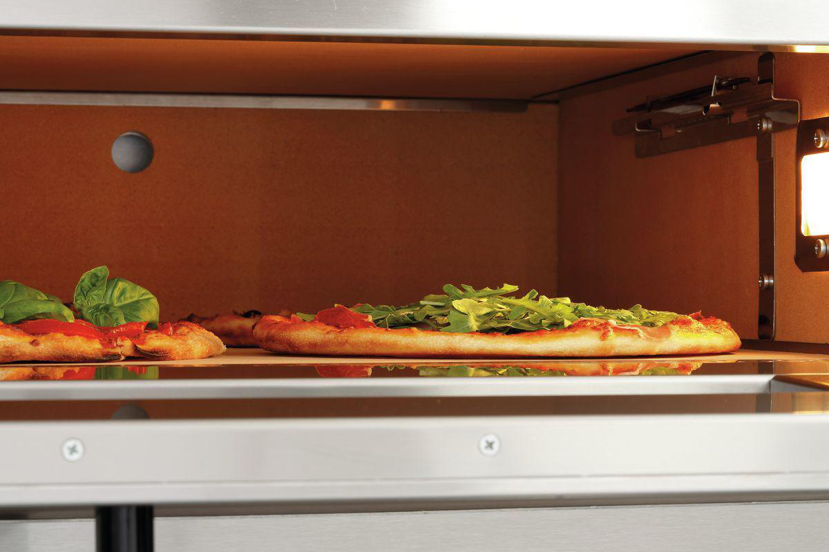 Im Ofen sind zwei Pizzen zu sehen. Eine hat Rucola, die andere Basilikum. Der Ofeninnenraum ist orange und das Äußere aus Edelstahl.