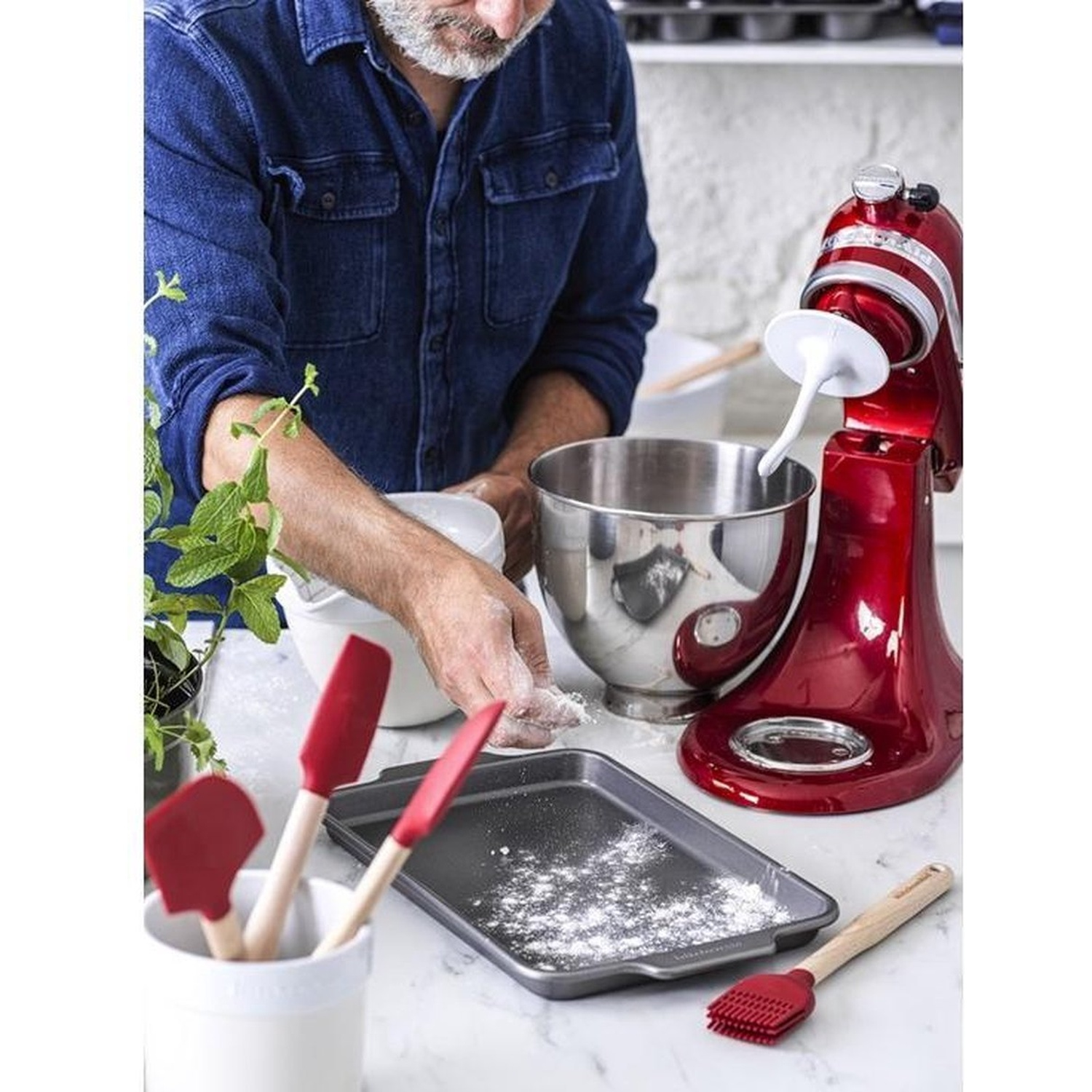 Un homme en jean prépare de la pâte dans une cuisine. Un batteur sur socle rouge, des spatules et une plaque à pâtisserie sont sur le comptoir.