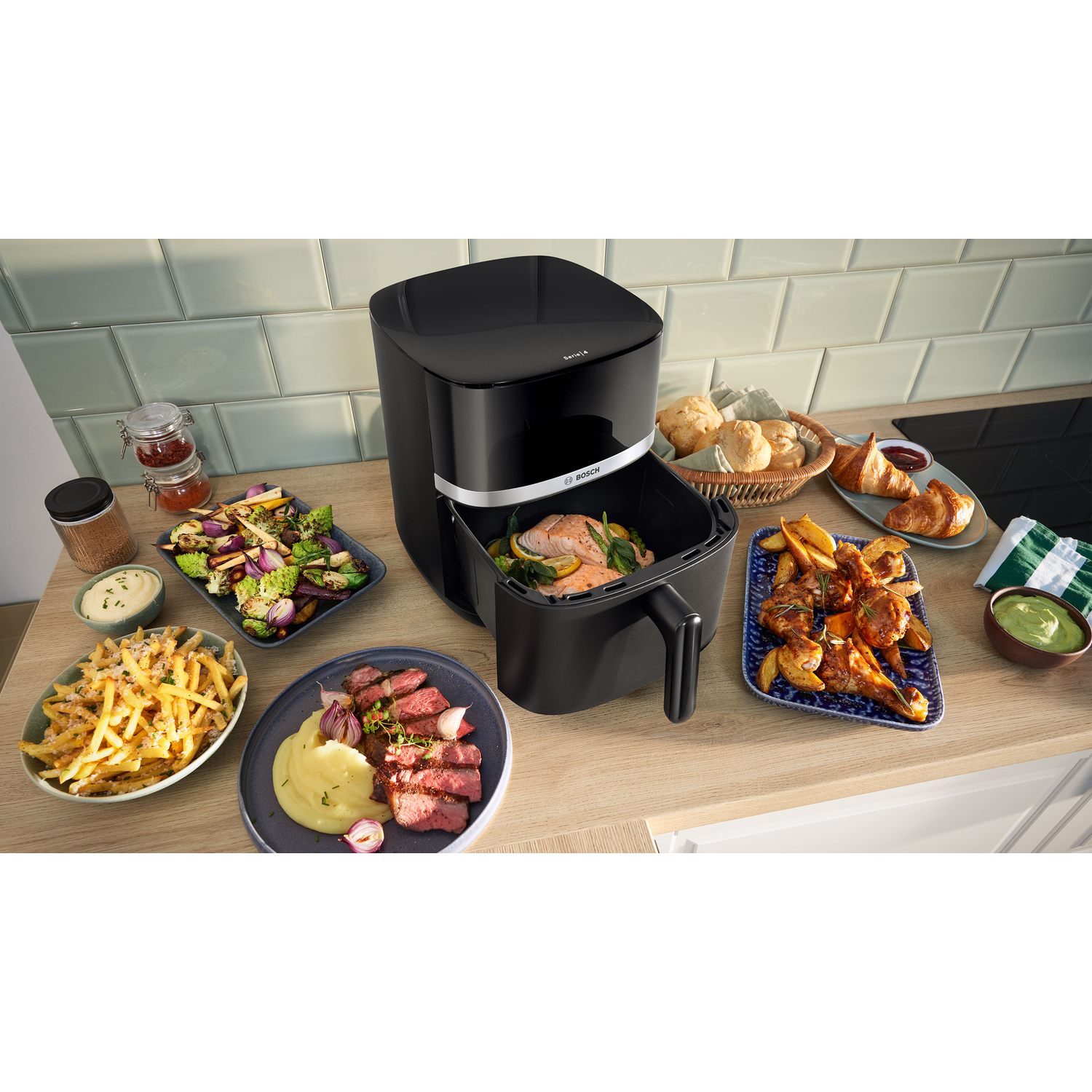 An air fryer with various dishes: fries, steak, vegetables, and chicken. The kitchen counter has various plates of food.