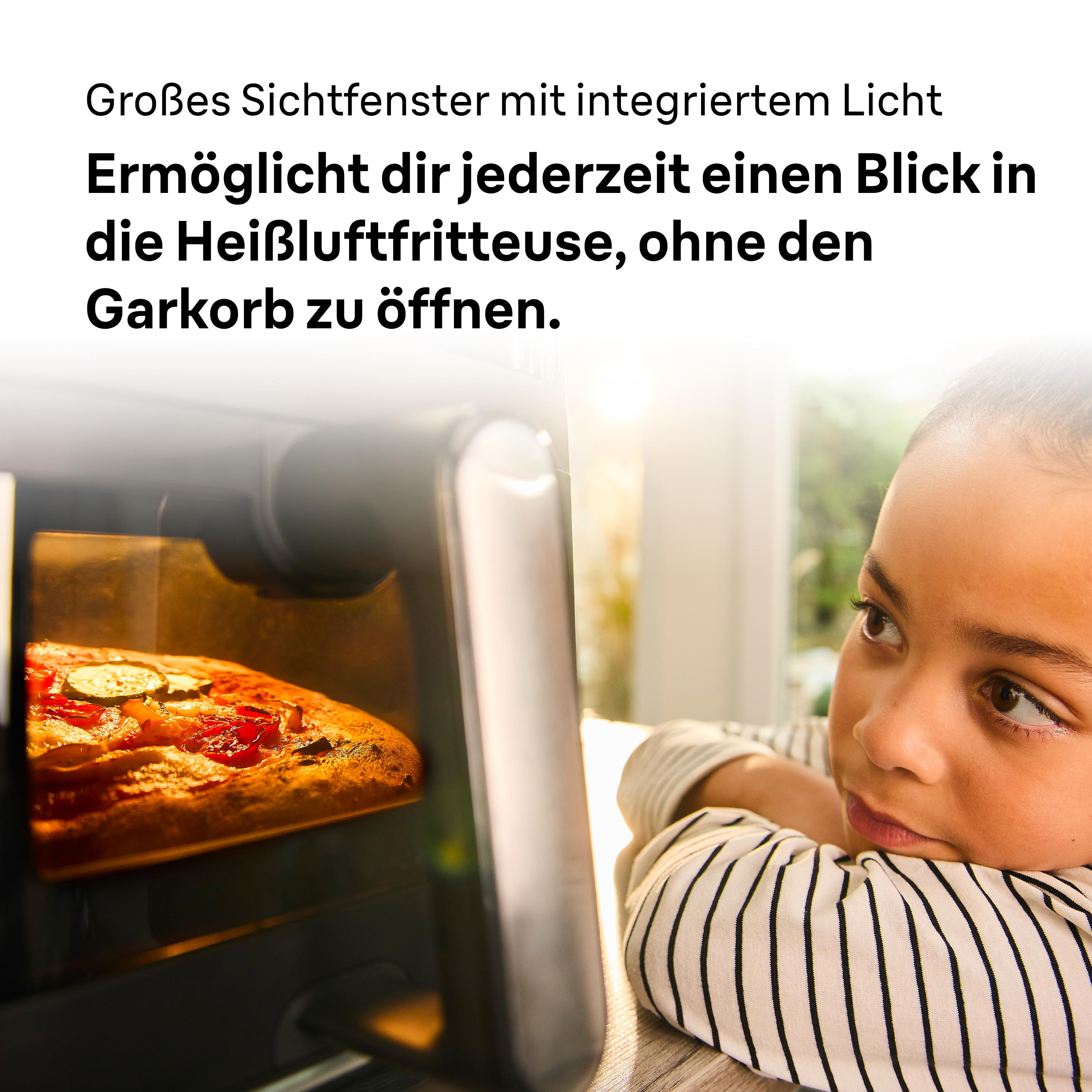 Ein Kind schaut auf eine Pizza, die in einer Heißluftfritteuse backt. Das Gerät hat ein Sichtfenster.