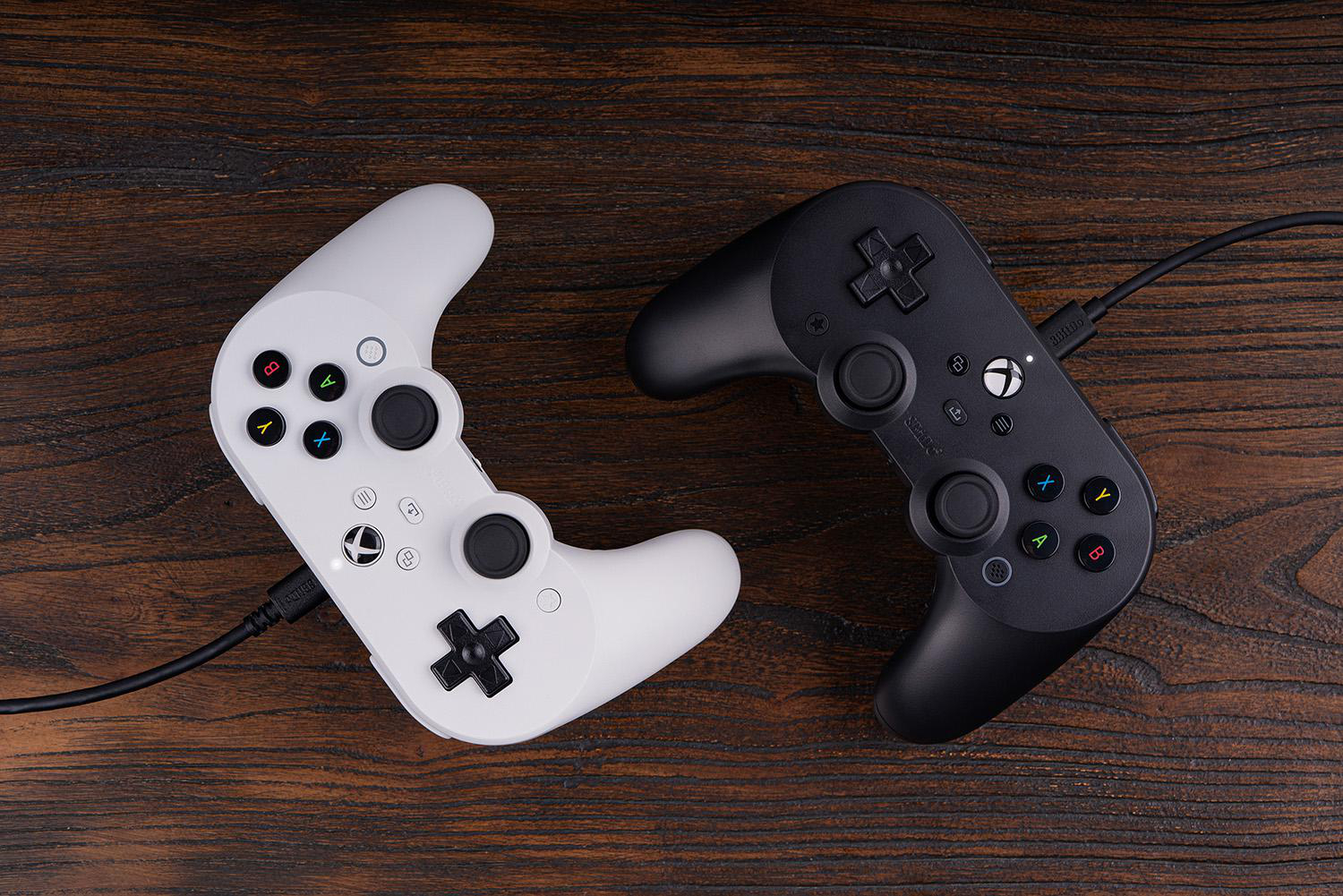 Zwei Gamecontroller, einer weiß und einer schwarz, liegen auf einer Holzoberfläche. Beide haben ein ähnliches Design und sind mit Kabeln verbunden.