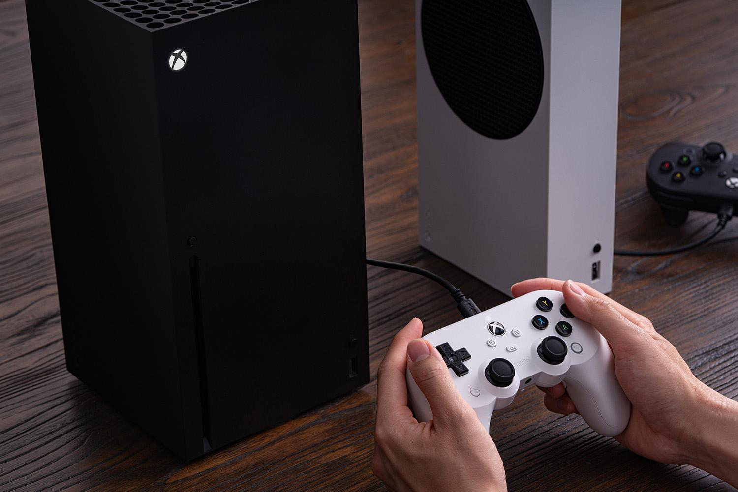 Xbox Series X und S Konsolen mit einem weißen Controller in den Händen. Ein zweiter Controller ist rechts. Dunkler Holztisch.