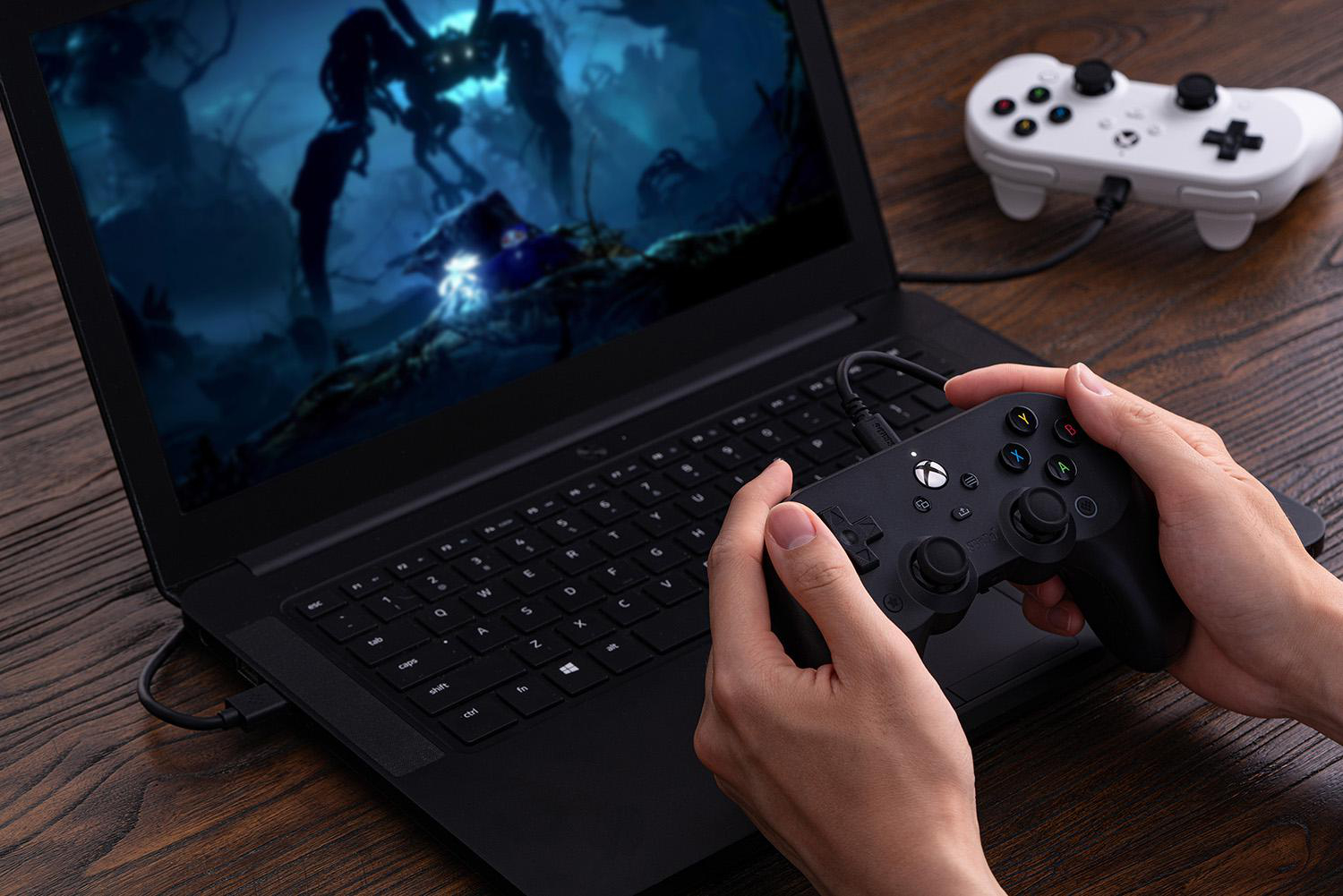 Eine Person hält einen schwarzen Xbox-Controller, der mit einem Laptop verbunden ist. Ein weißer Controller liegt neben dem Laptop.