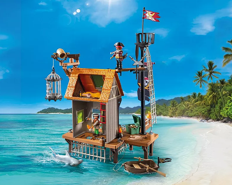 Ein Piratenschiff-Spielzeugset am Strand. Ein Hai schwimmt in der Nähe des Docks. Palmen und blauer Himmel im Hintergrund.