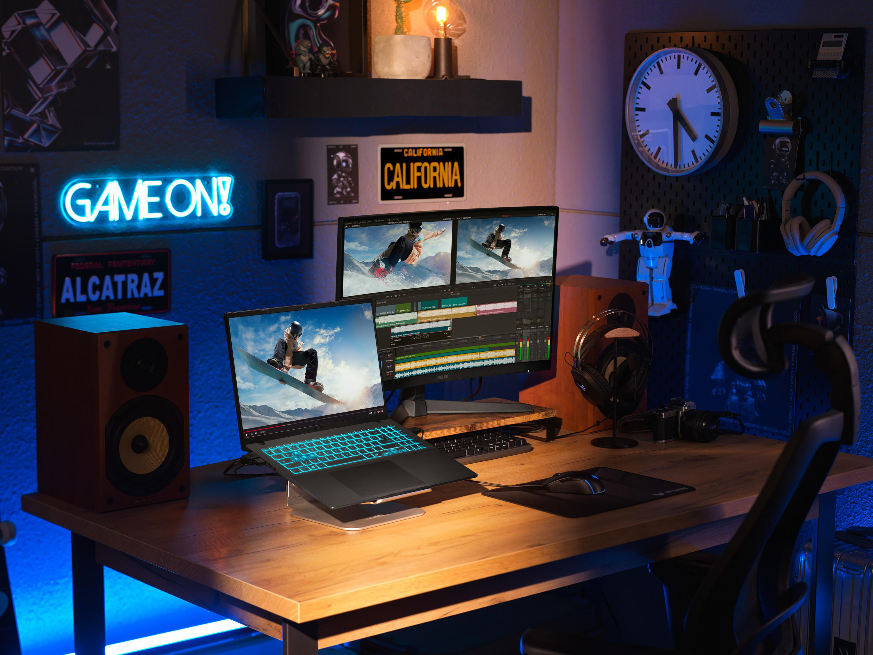 Gaming-Setup mit Laptop, Monitoren mit Snowboarder, Neon-Schild, Lautsprechern und Uhr auf einem Holzschreibtisch.