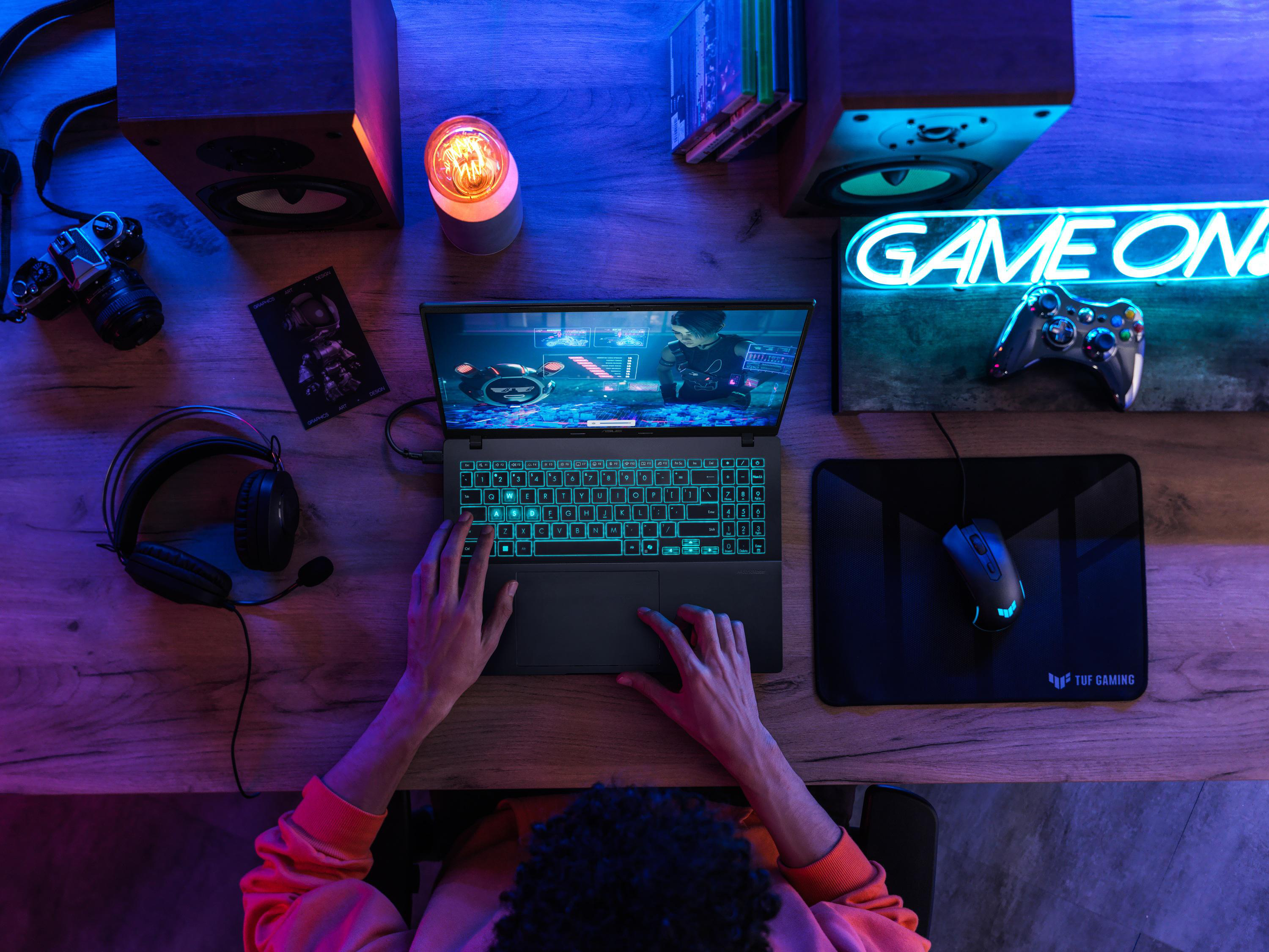 Eine Person spielt auf einem Laptop. Das Gaming-Setup umfasst Lautsprecher, Kopfhörer und einen Gamecontroller. Das Neon-Schild sagt GAME ON.