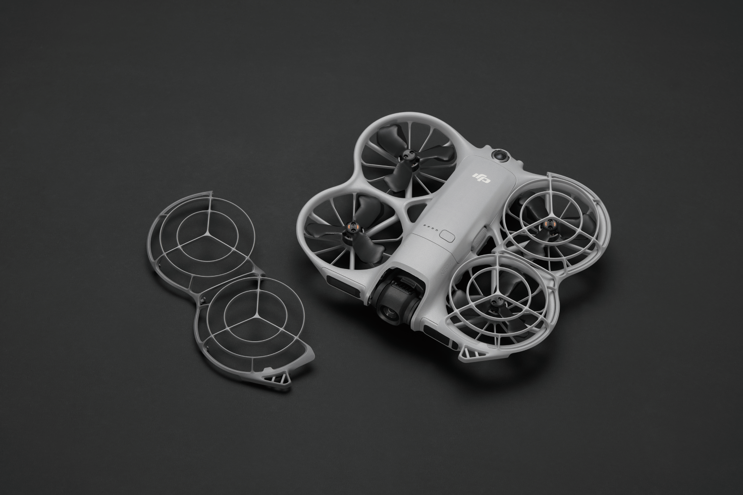 Un dron gris con cuatro hélices y una jaula protectora está sobre una superficie negra. También hay una jaula separada.
