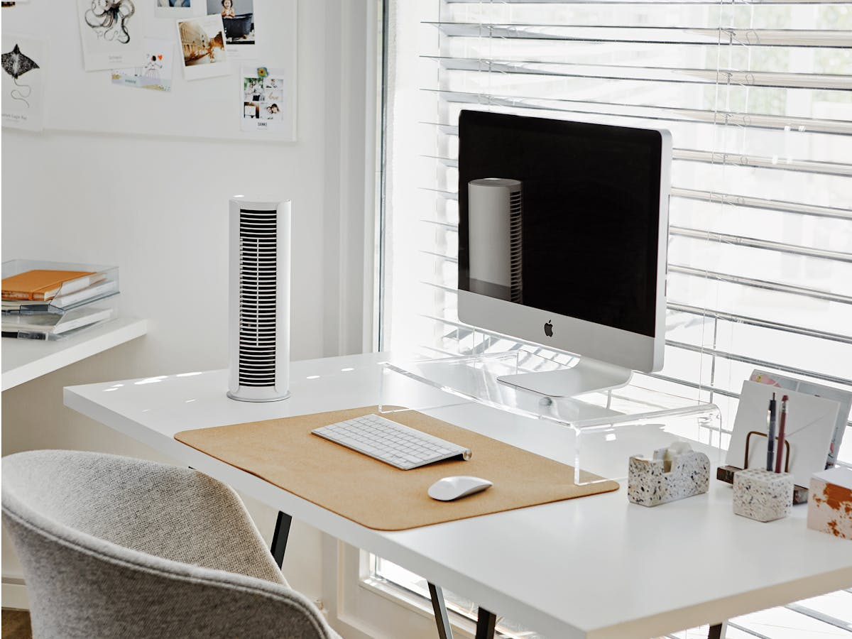 Una scrivania moderna con computer, tastiera, mouse, ventilatore e decorazioni. La scrivania è bianca, la sedia grigia.