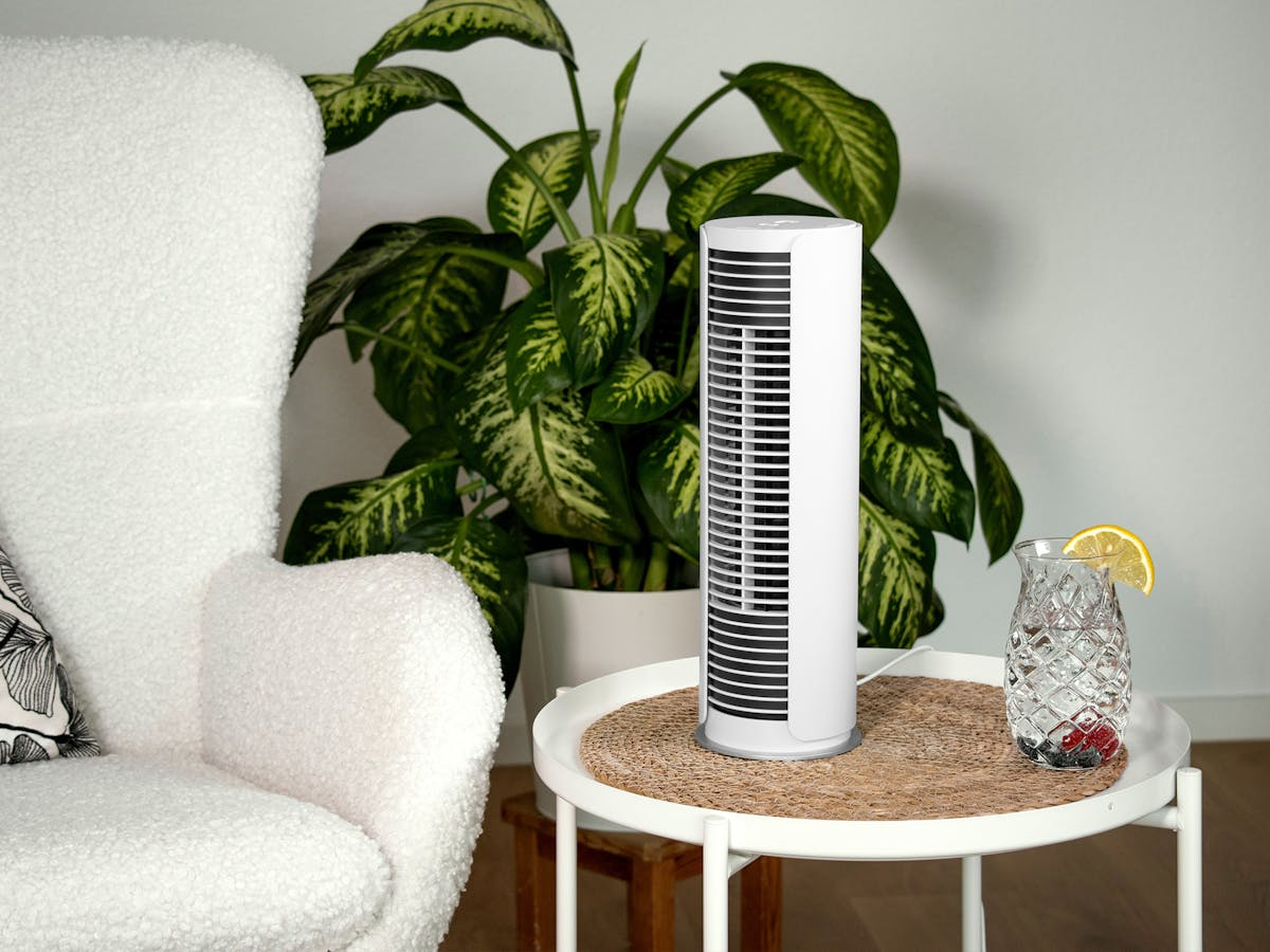 Un ventilatore a torre bianco su un tavolo con una bevanda, una pianta e una sedia. La sedia è bianca, la pianta ha grandi foglie verdi.
