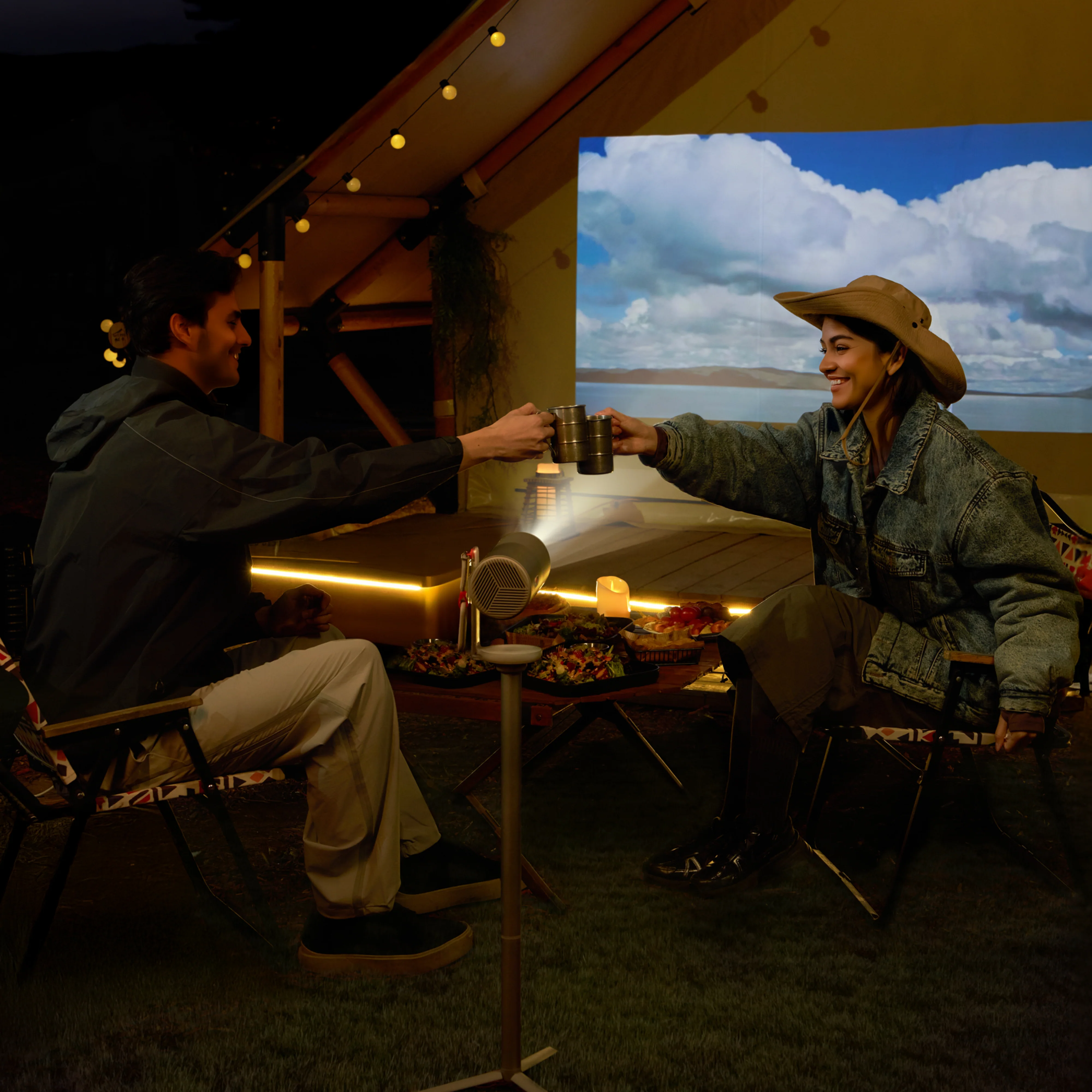 Deux personnes trinquent avec des mugs dans une scène de camping. Un projecteur affiche un paysage sur un écran. De la nourriture est sur la table.
